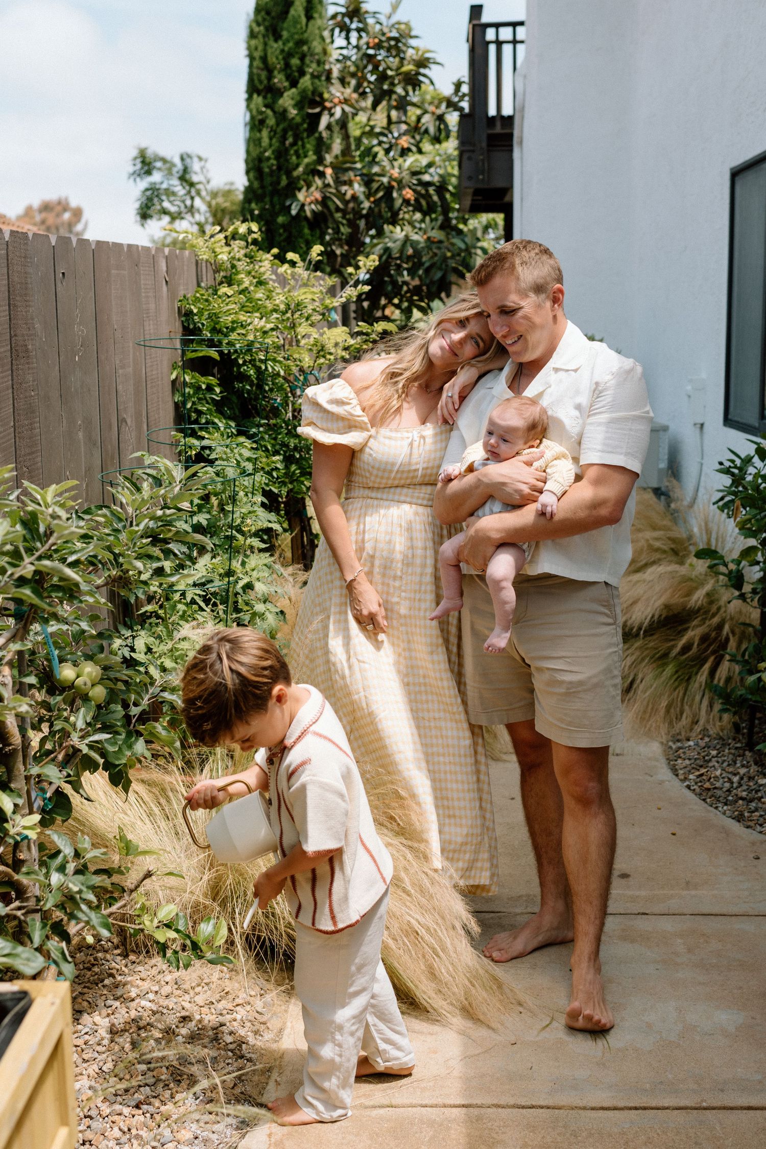 Young family with toddler and newborn baby posing in lush backyard garden.
