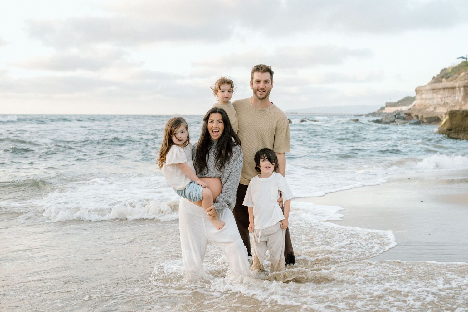 Happy family of five posing together on a sandy beach with ocean waves, rocky cliffs in the background.