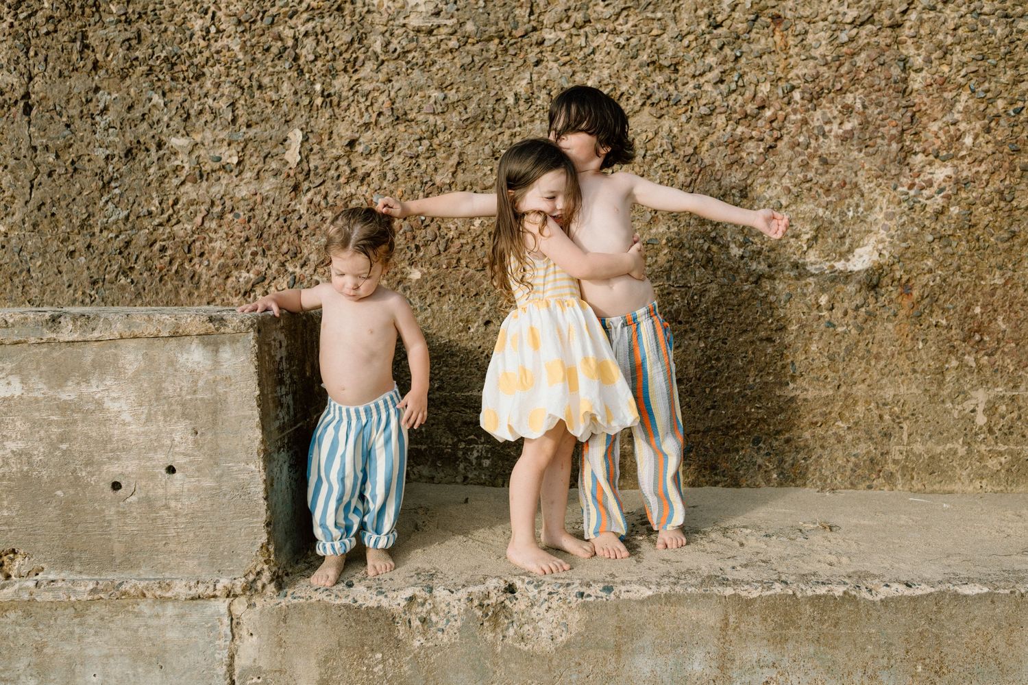 Three young children in colorful striped shorts play and hug together against a textured stone wall.