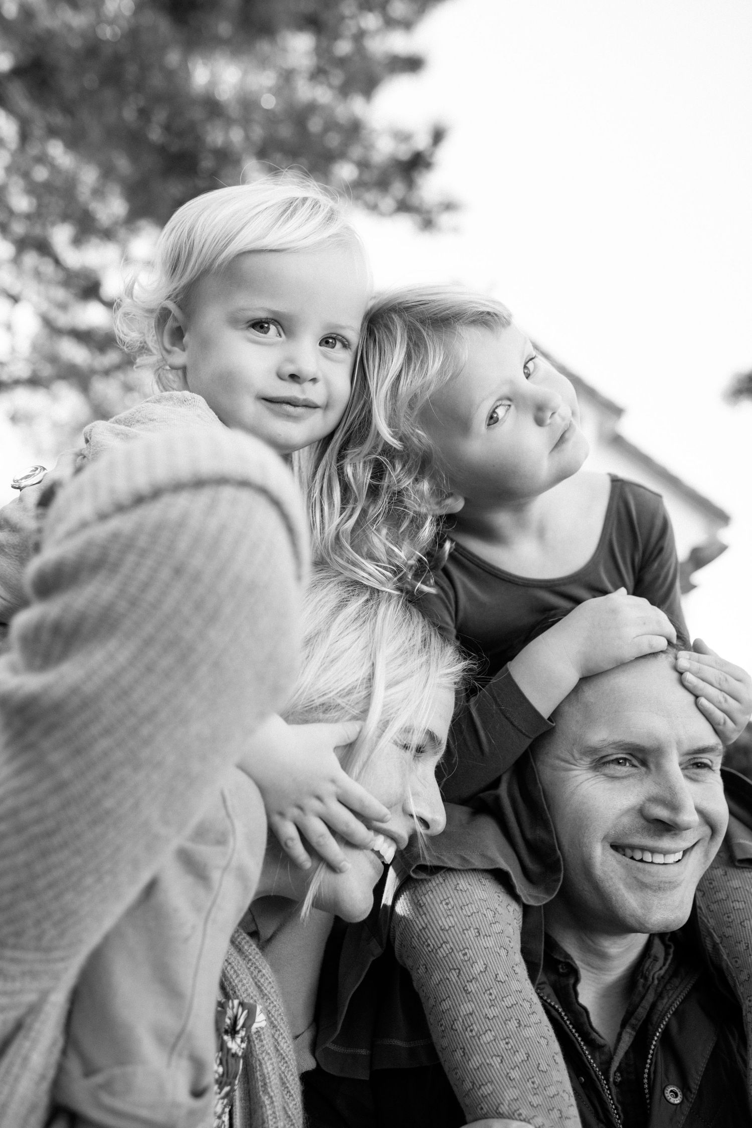 Black and white photo of a smiling father with two young blonde children sitting on his shoulders outdoors.