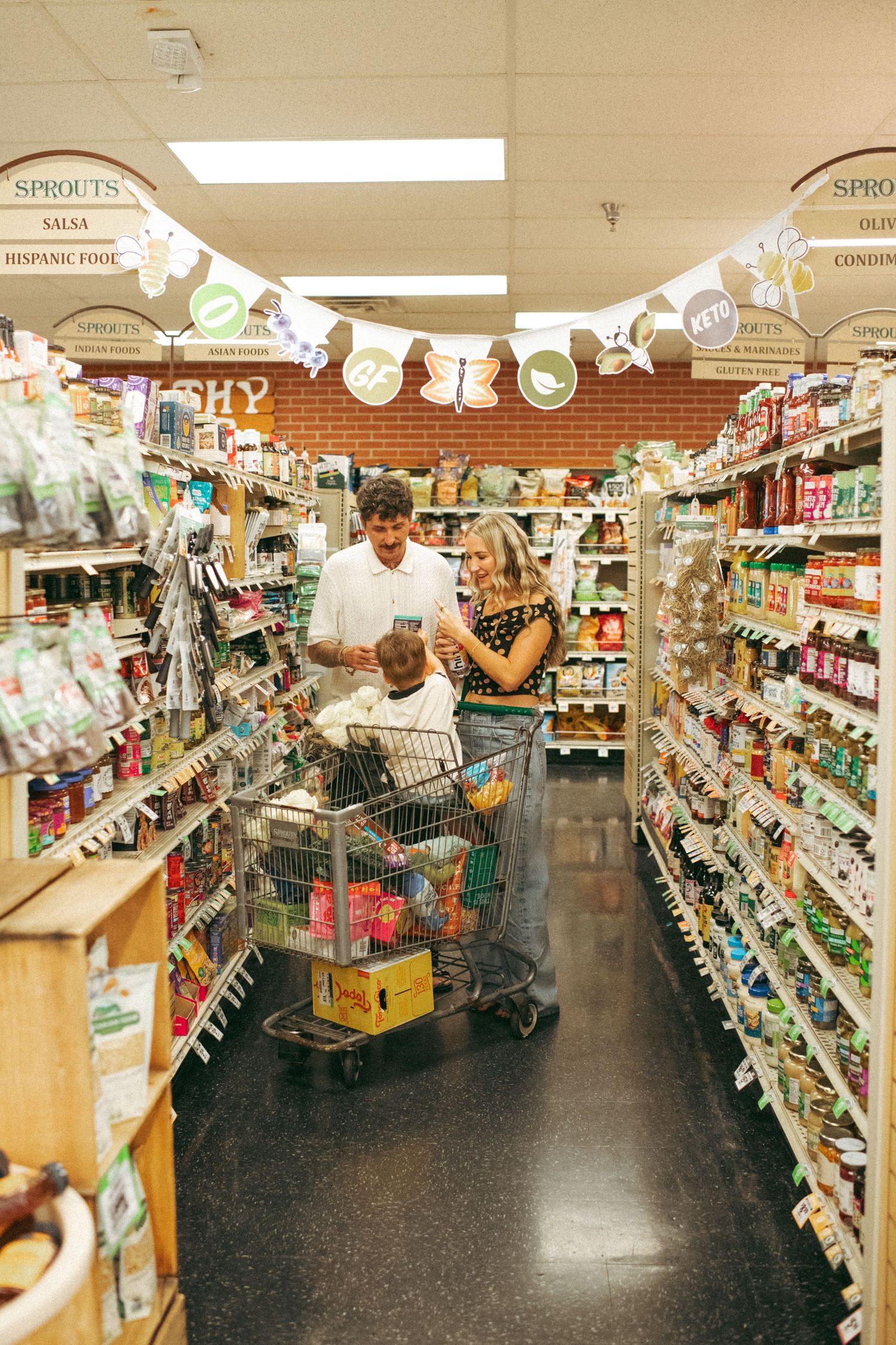 Family shopping in Sprouts grocery store aisle, parents pushing cart with child inside, browsing products.