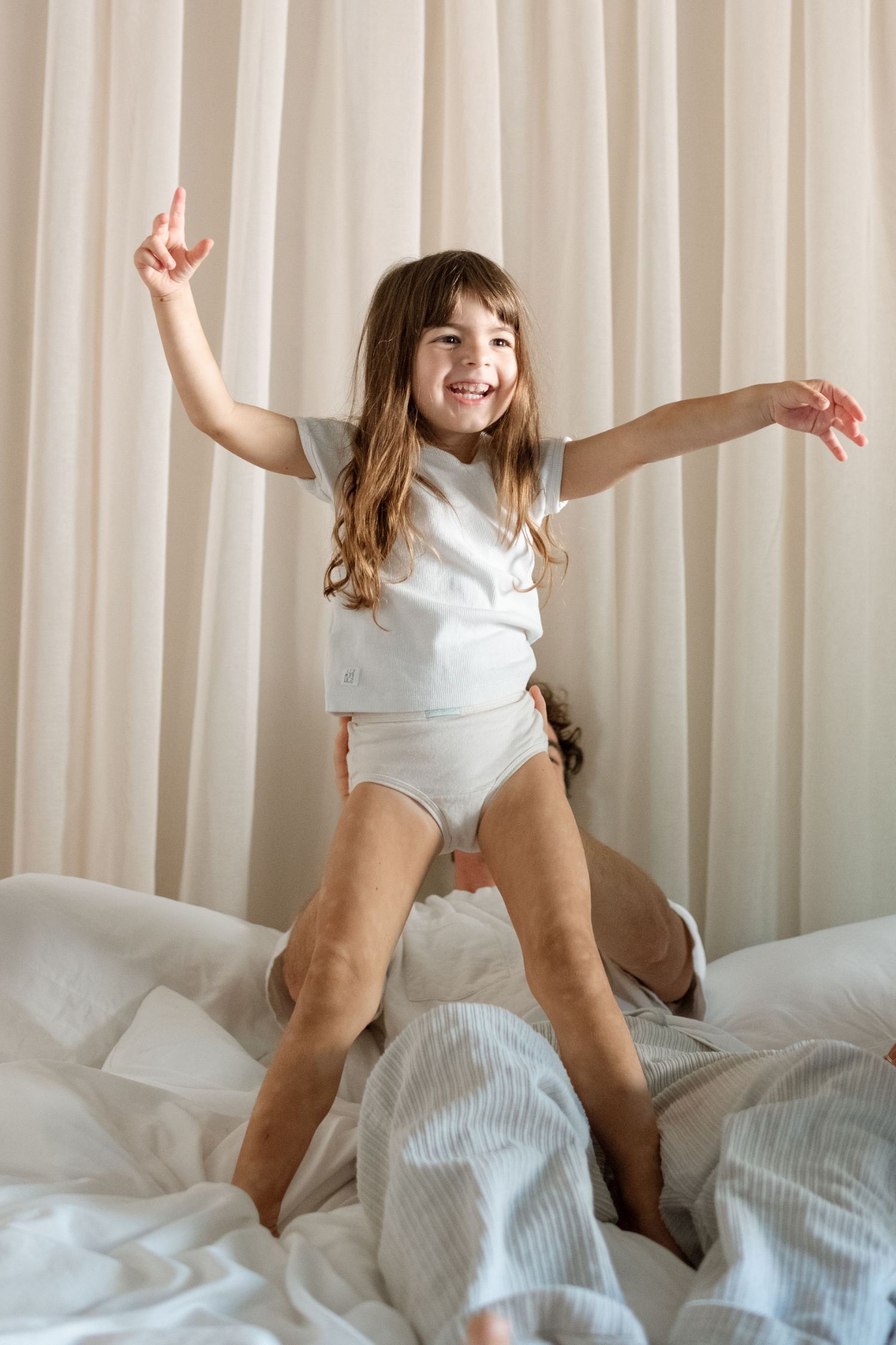 Happy young girl in white pajamas jumping on a bed with arms outstretched, smiling joyfully.