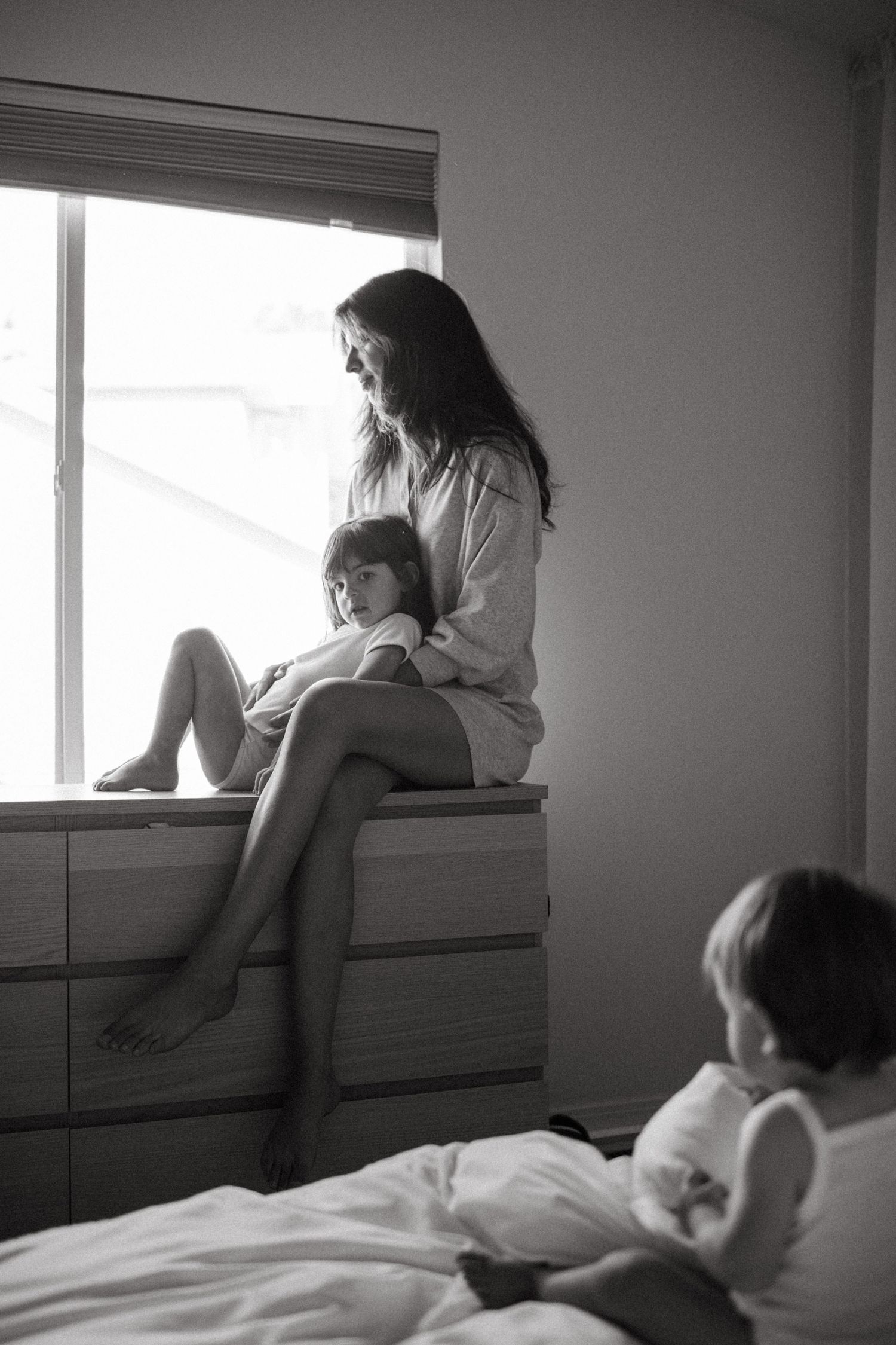 Black and white photo of a mother and child sitting on a dresser by a window while another child watches from a bed.