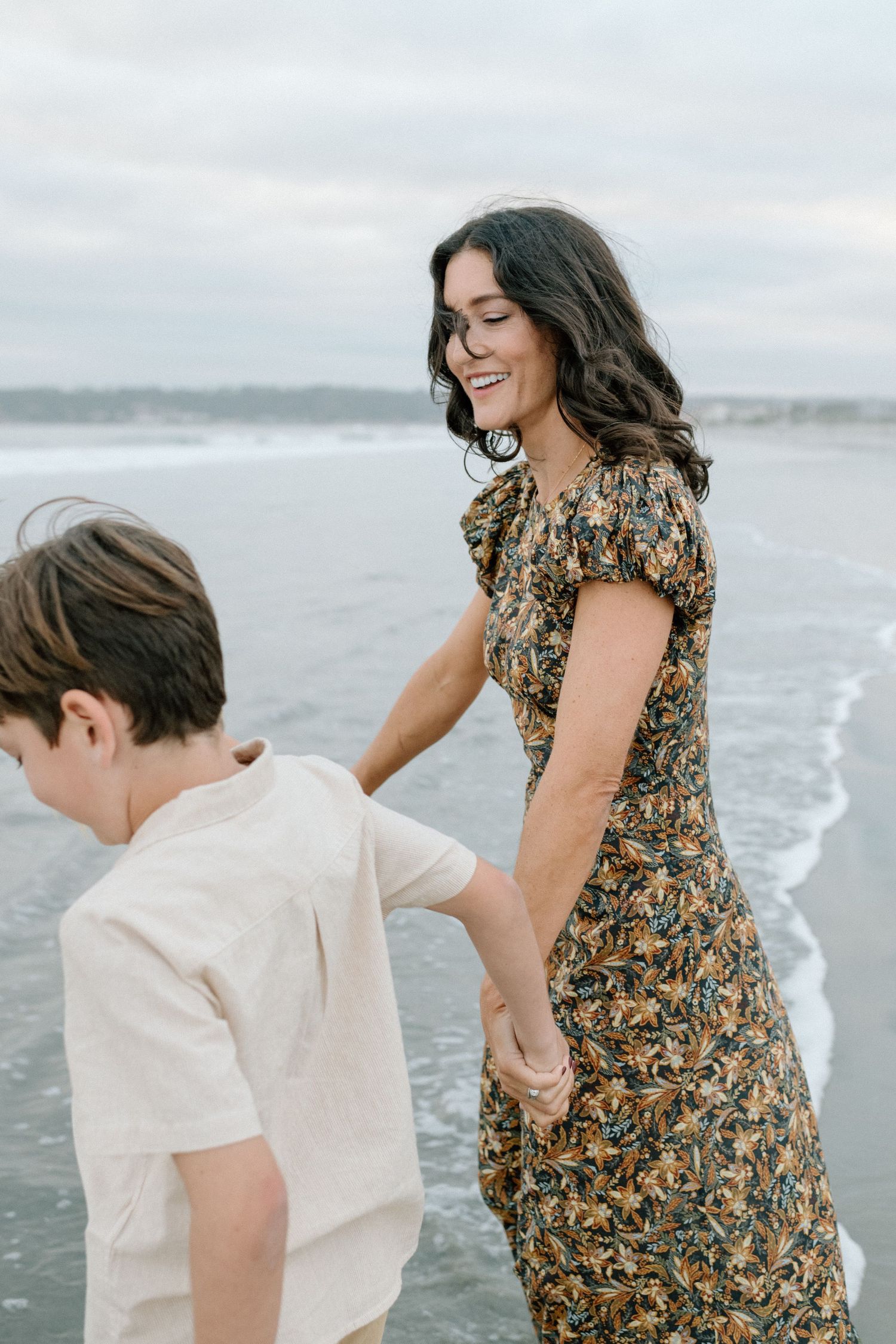 A smiling mother in a floral dress holds her sons hand while walking along a cloudy beach shoreline.