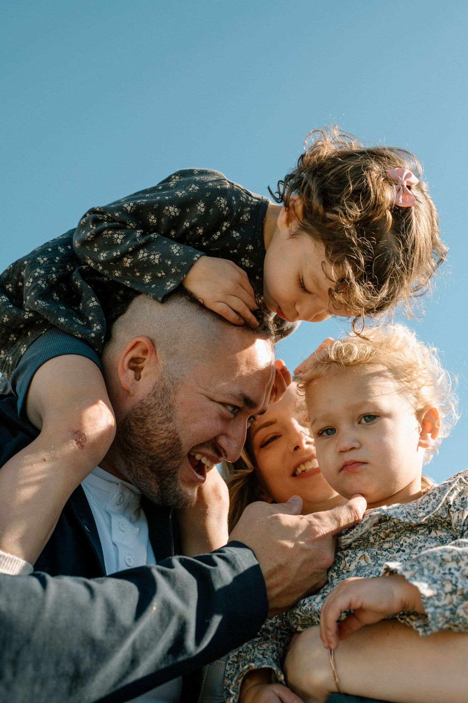 Father and mother hugging two young curly-haired children under blue sky.