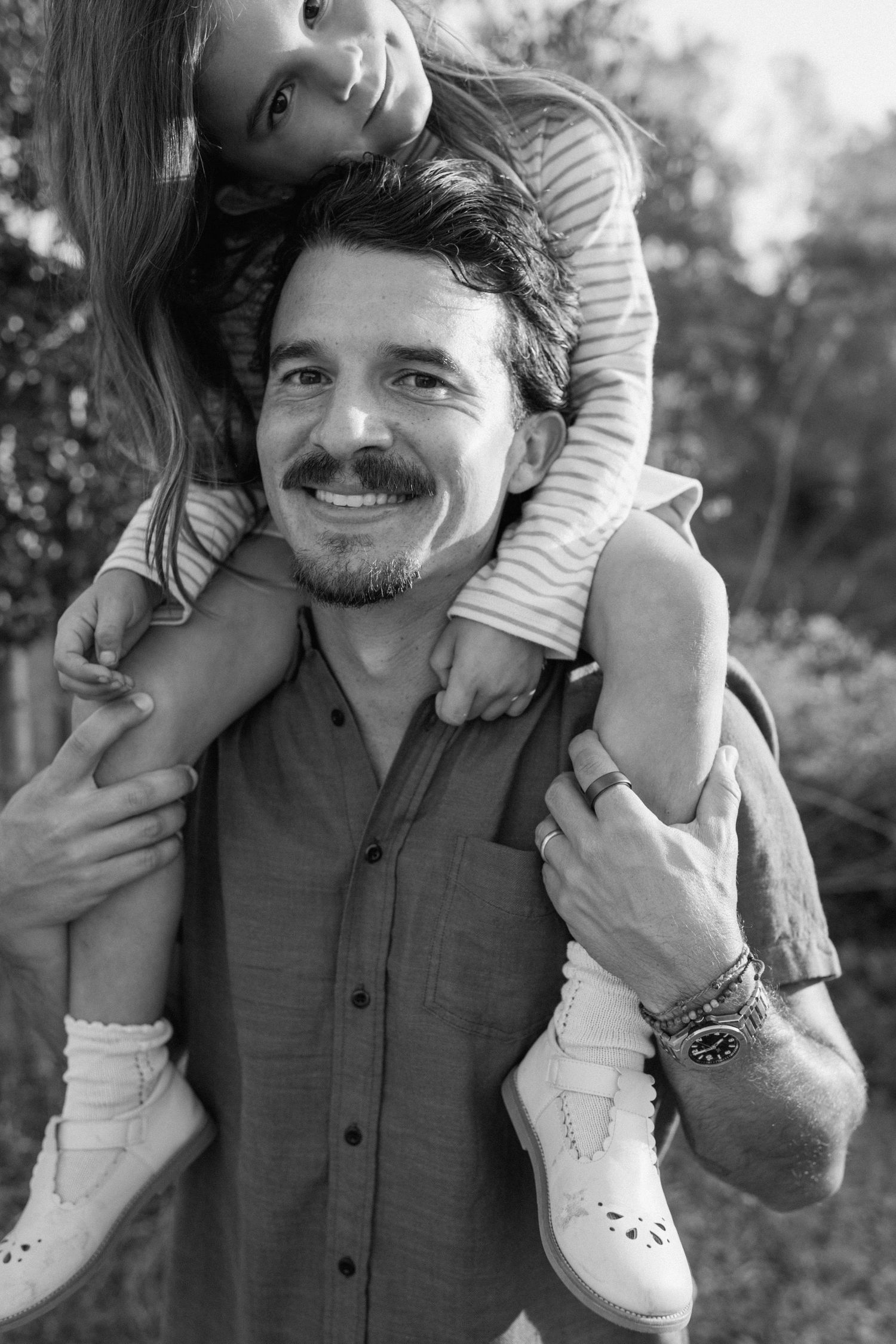 Black and white portrait of smiling father carrying young daughter on his shoulders outdoors.