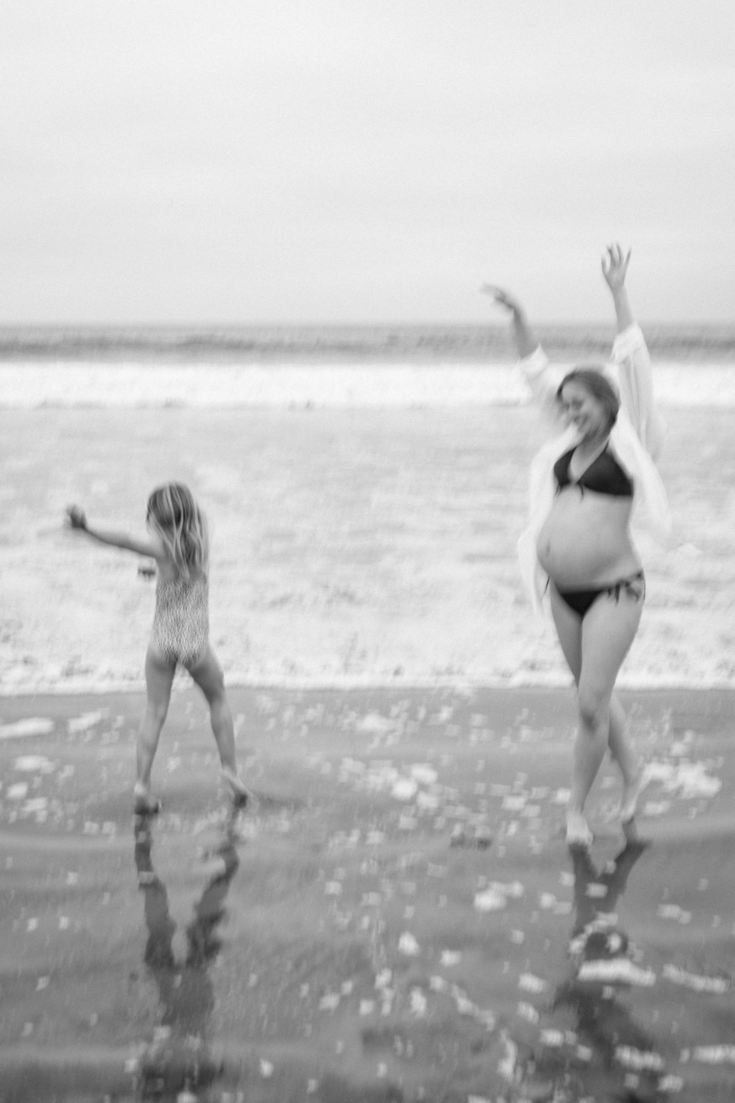 Black and white blurred image of pregnant woman and young girl dancing joyfully at waters edge.