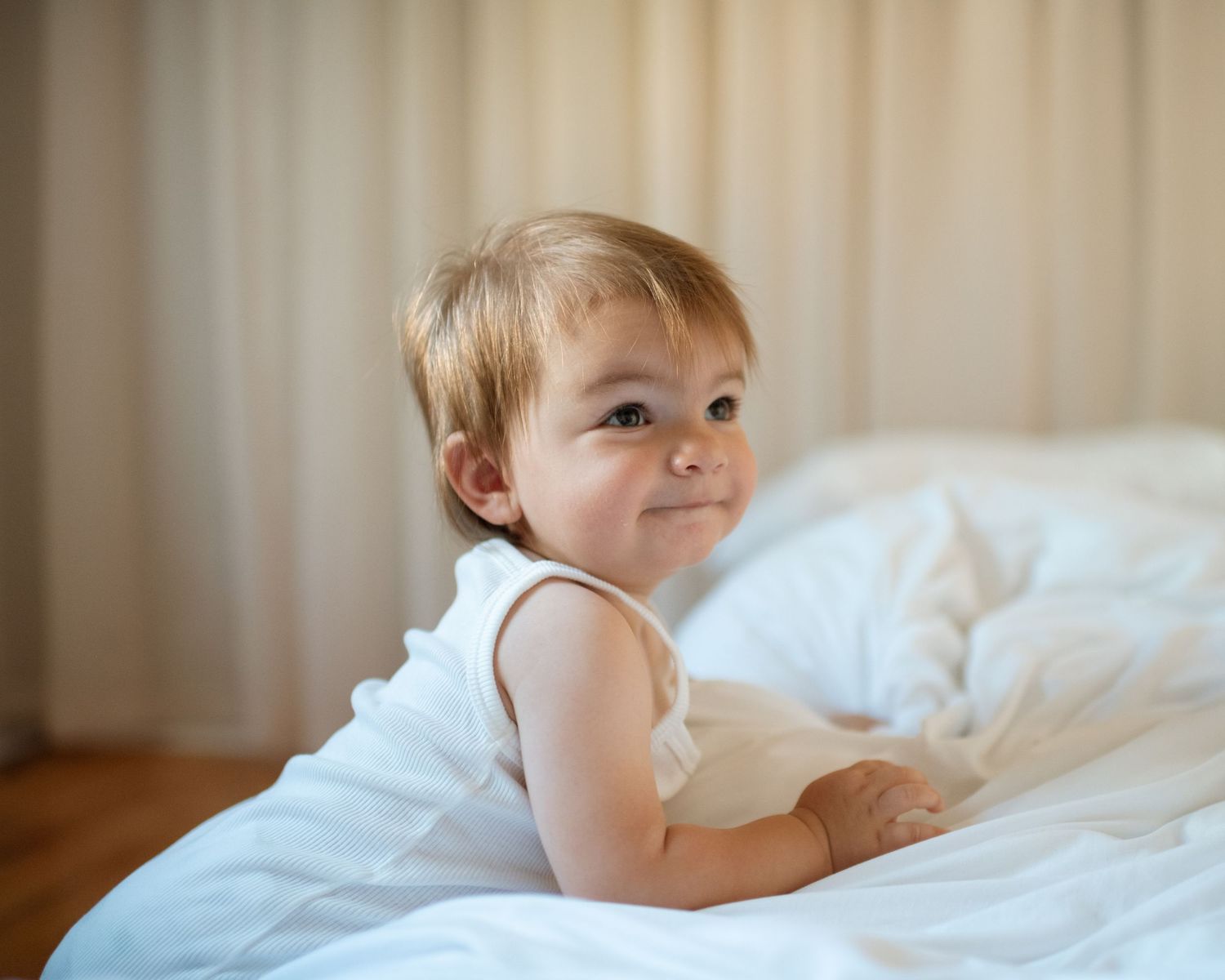 Cute toddler in white sleeveless outfit smiling on a white bed with soft natural light.