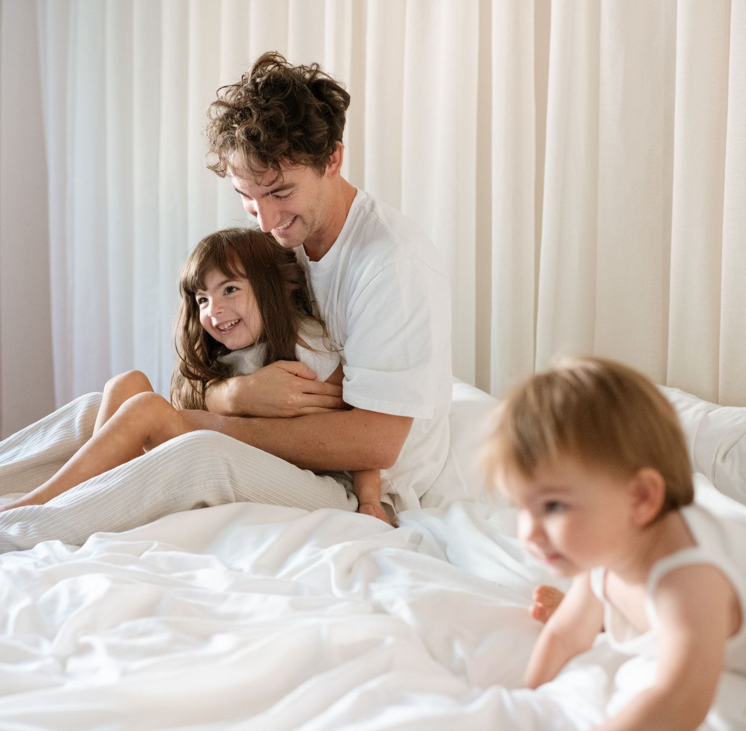 Father smiling and hugging young daughter on white bed while toddler plays in foreground.