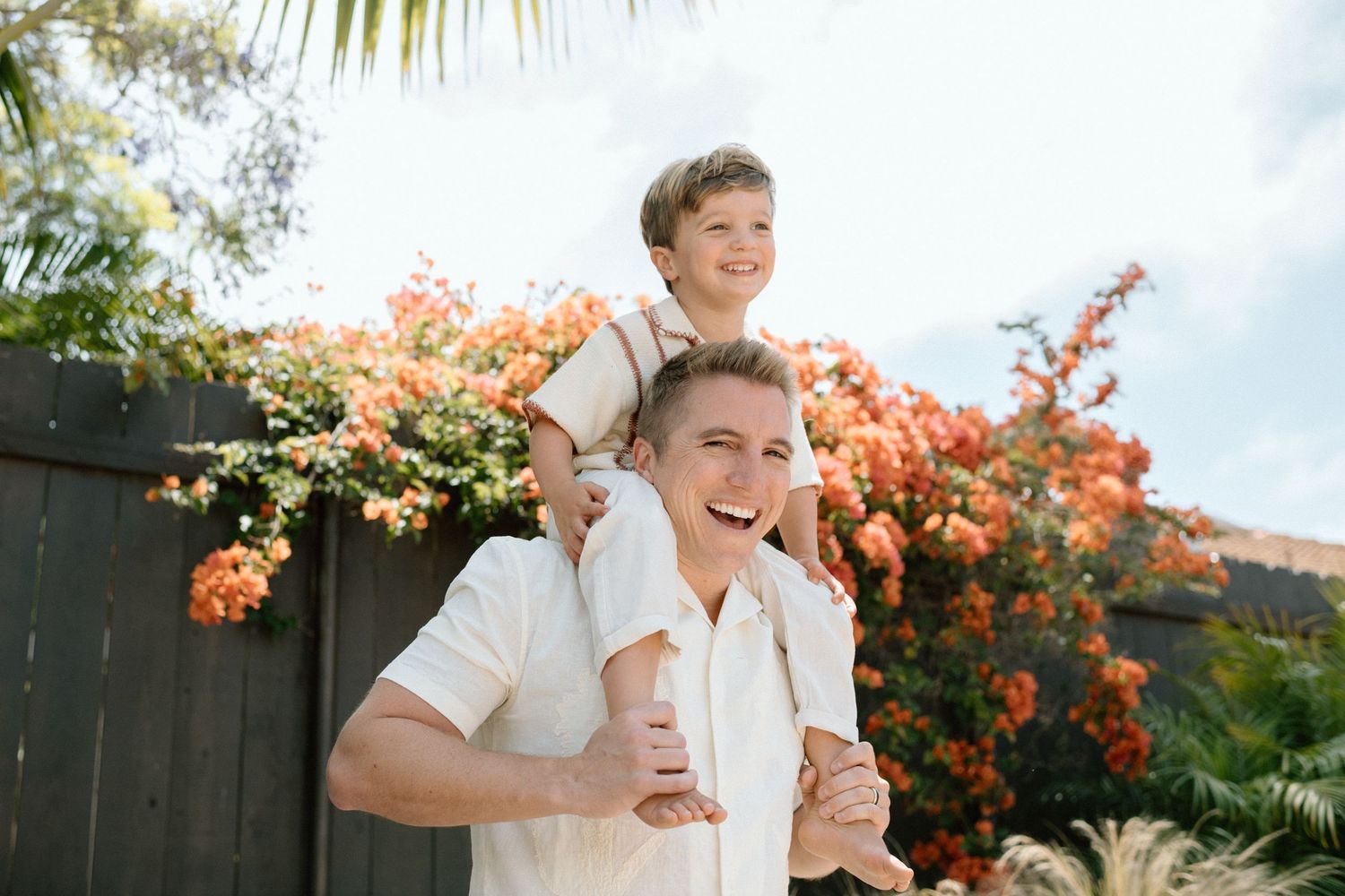 Laughing father carrying young son on shoulders outdoors near colorful orange bougainvillea flowers.