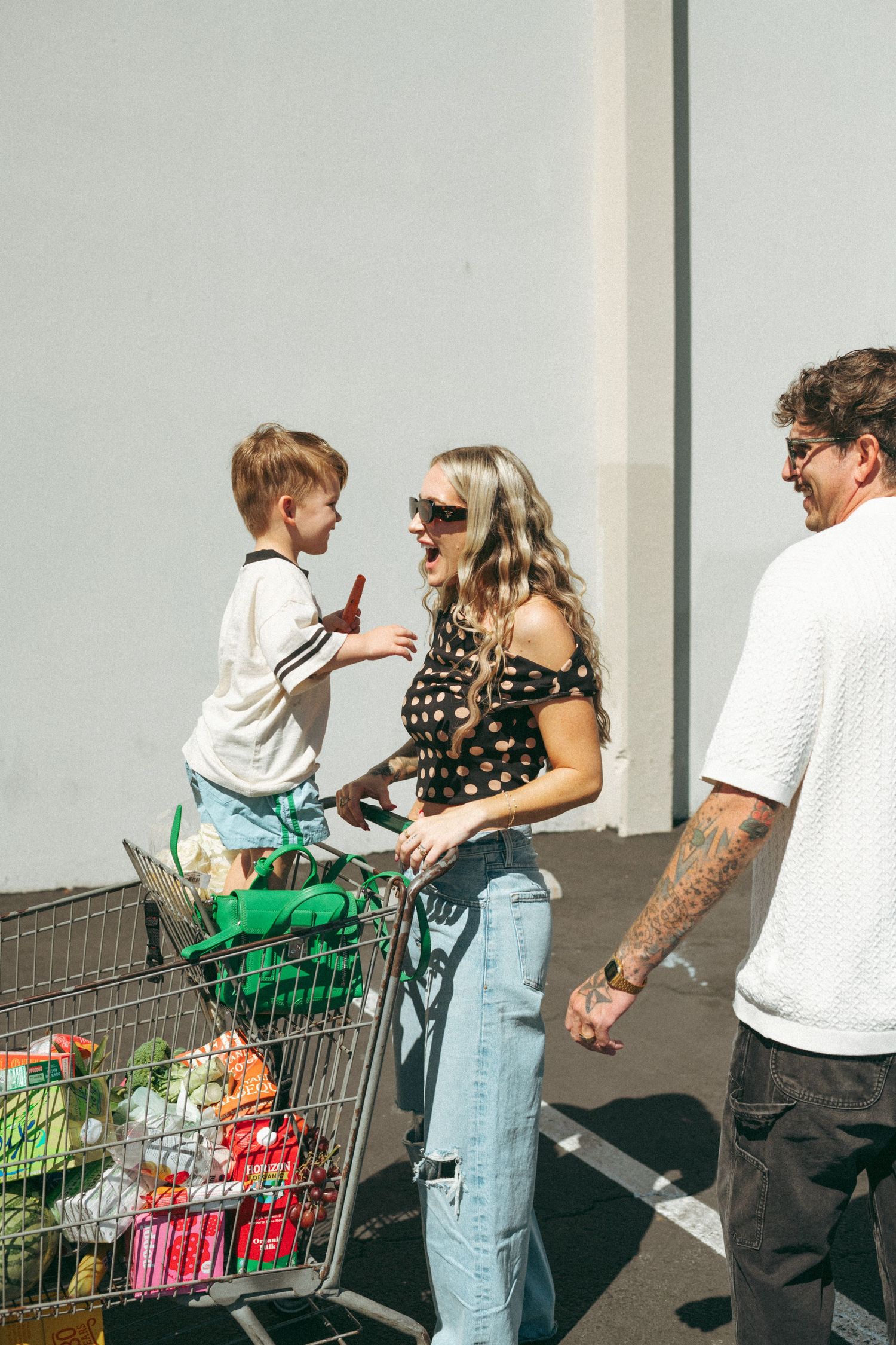 Family with tattooed dad, blonde mom in polka dot top, and young son sitting in grocery cart filled with fresh produce.