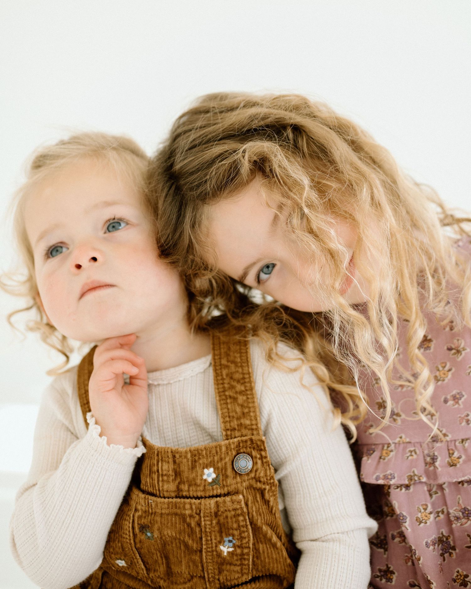 Two young blonde sisters cuddle close together, older girl leaning her head against younger sibling.