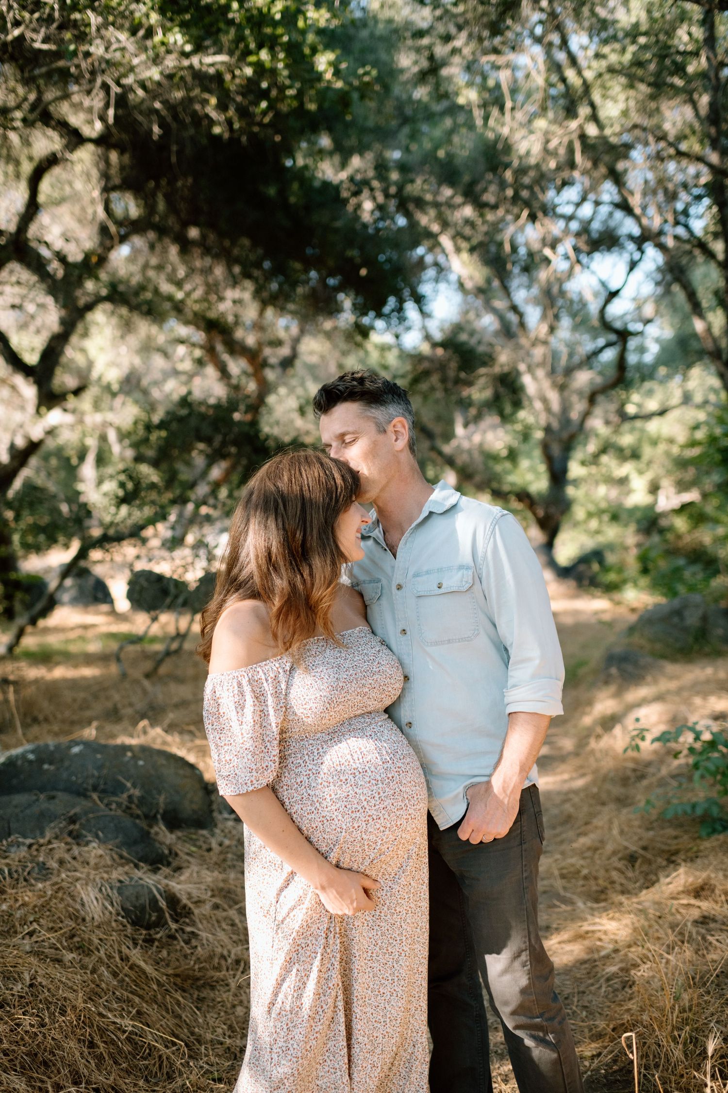 Couple sharing tender moment outdoors, pregnant woman in floral dress embraced by partner among oak trees.