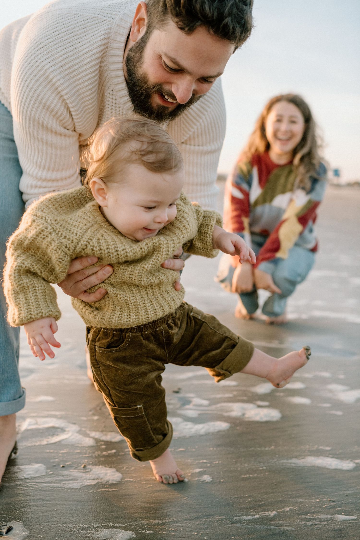 Parents help their smiling baby take first steps on a wet sandy beach at sunset.