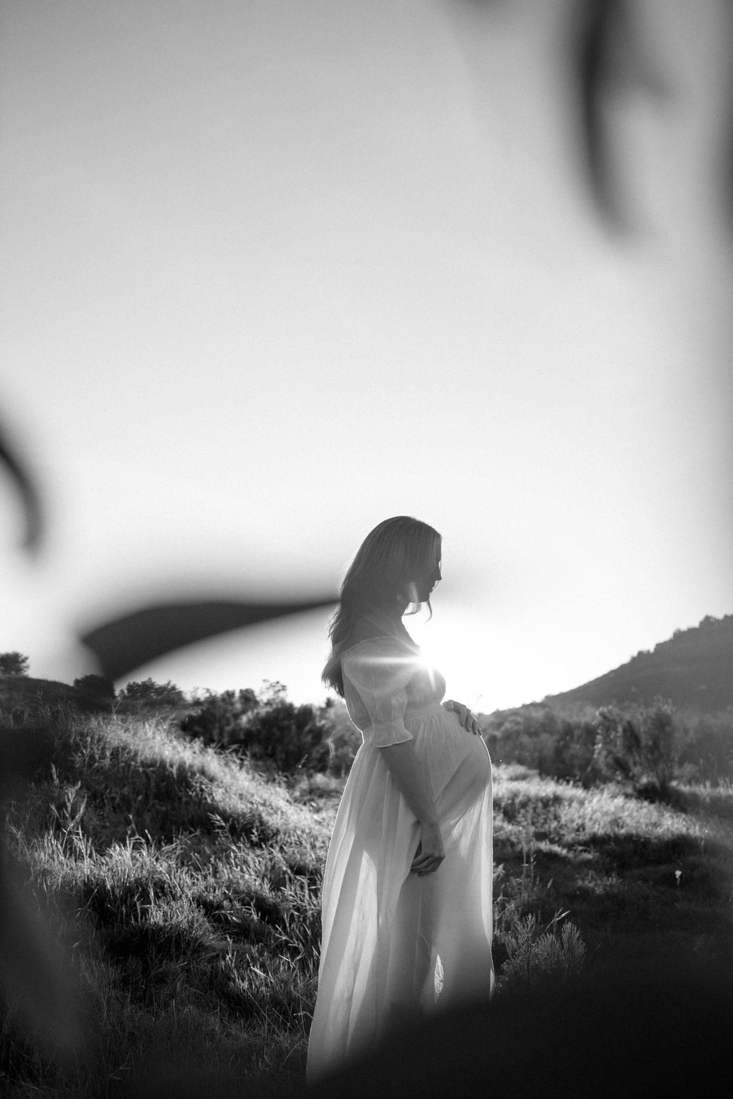 Black and white silhouette of pregnant woman in white dress standing in sunlit outdoor landscape.