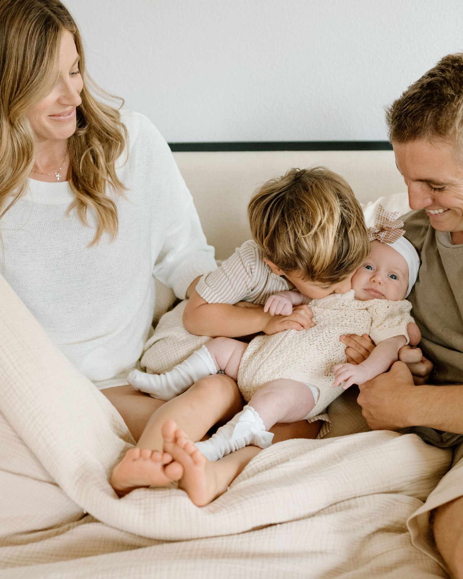 Happy family with parents, toddler boy kissing newborn baby girl sitting on a cozy bed.