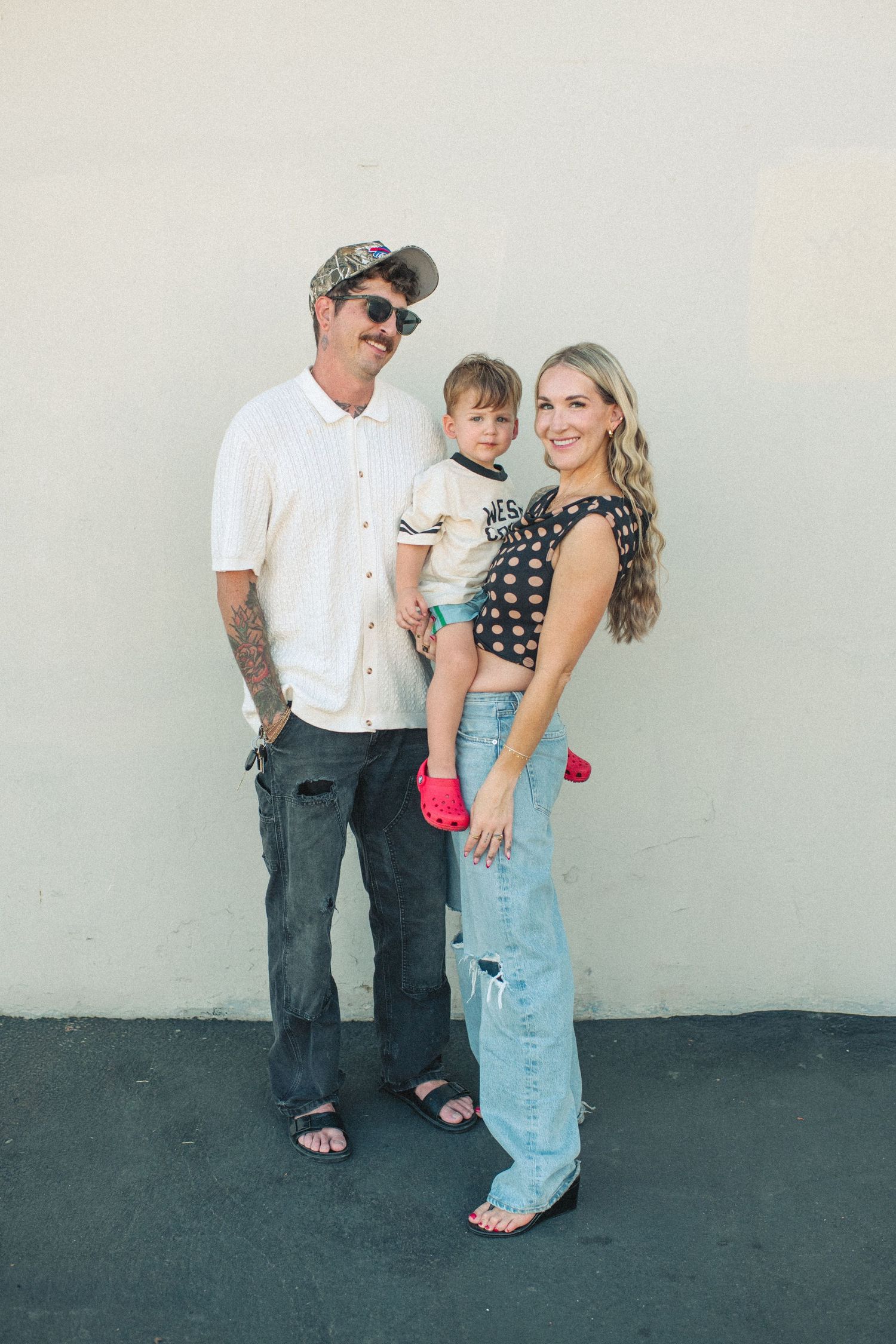 A tattooed man and blonde woman pose with their toddler against a white wall in casual stylish outfits.