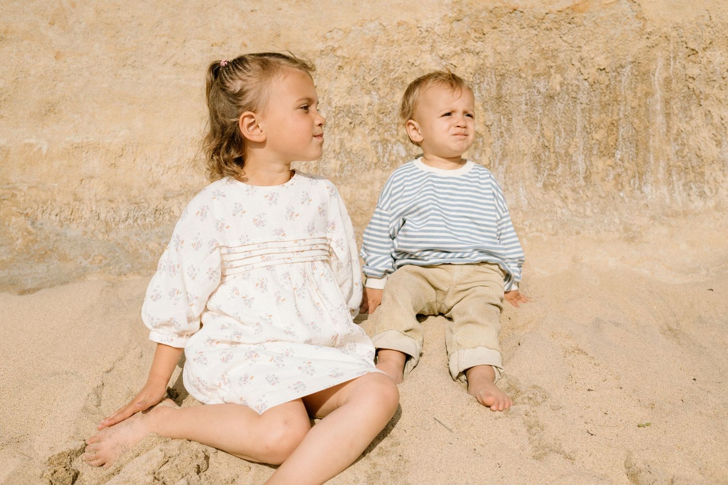 Two young children sit together on sandy beach, girl in floral dress and boy in striped shirt.