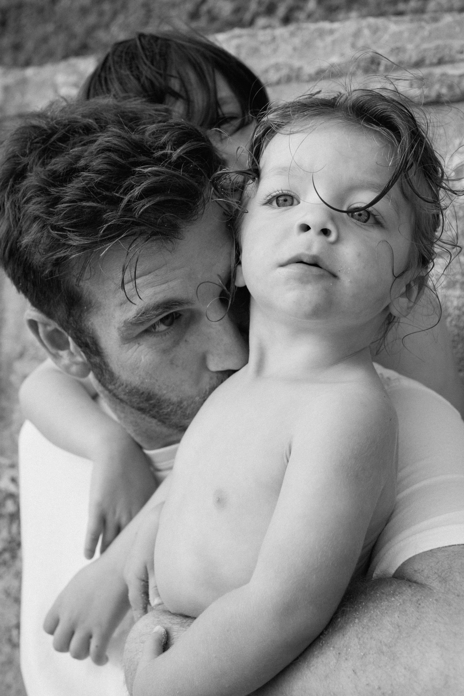 Black and white portrait of father holding curly-haired blue-eyed toddler with mother behind.