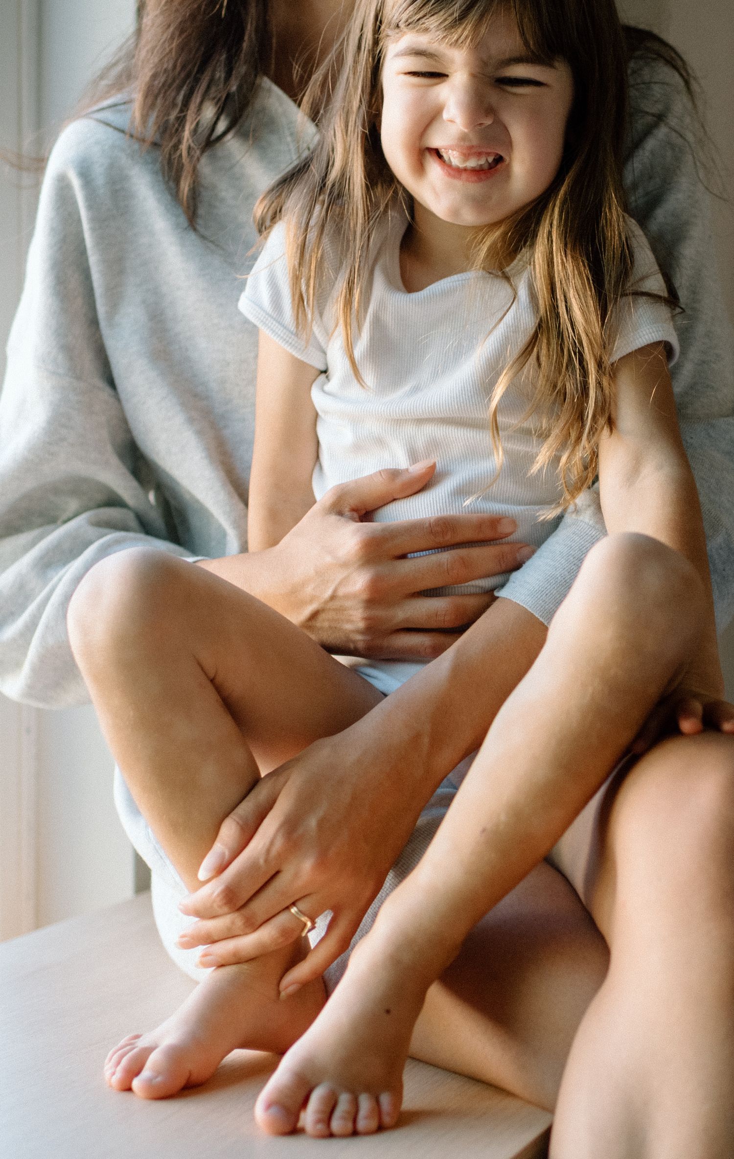A laughing young girl sits cross-legged on a woman's lap while being tickled, both barefoot indoors.