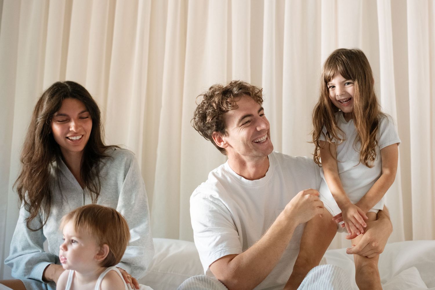 Happy family with two young children playing and laughing together on a white bed near sheer curtains.
