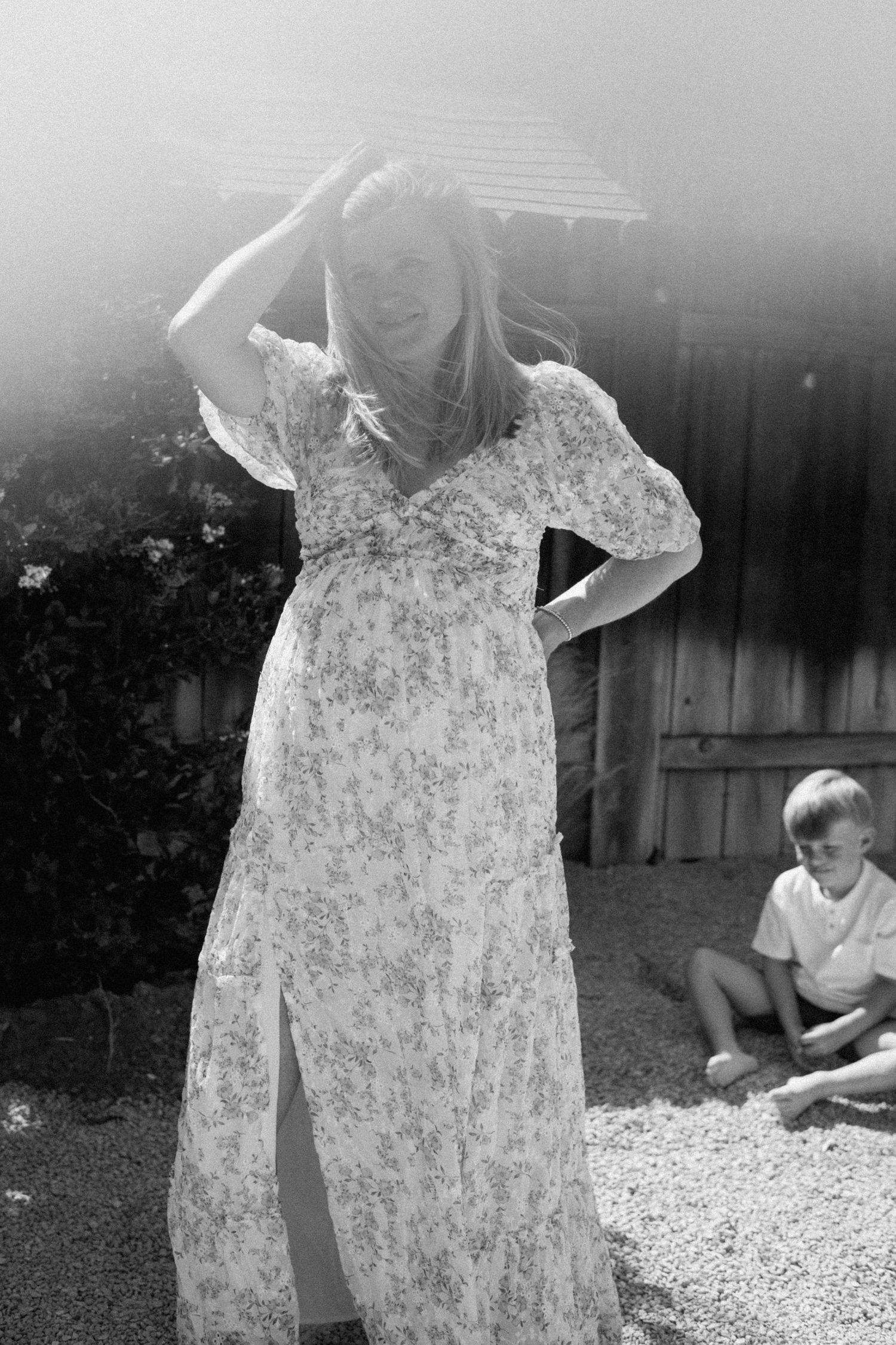 Black and white photo of a pregnant woman in a floral dress shielding her face outdoors on a sunny day.