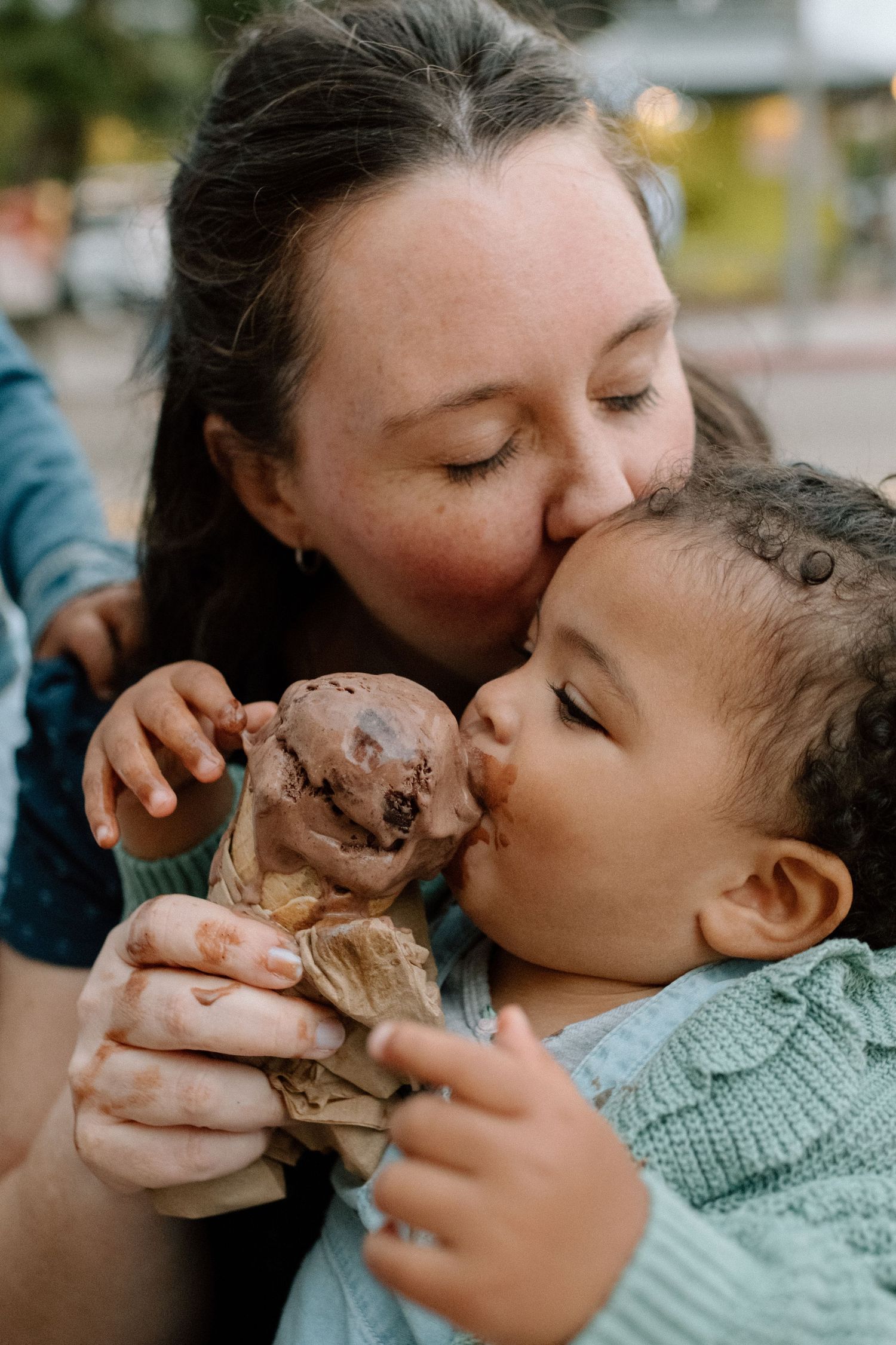 Mother kissing toddler on the head while child eats a melting chocolate ice cream cone.
