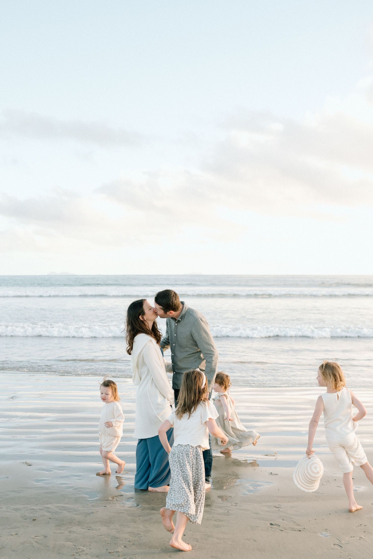 Parents sharing a kiss on the beach while their five children play in the waves.