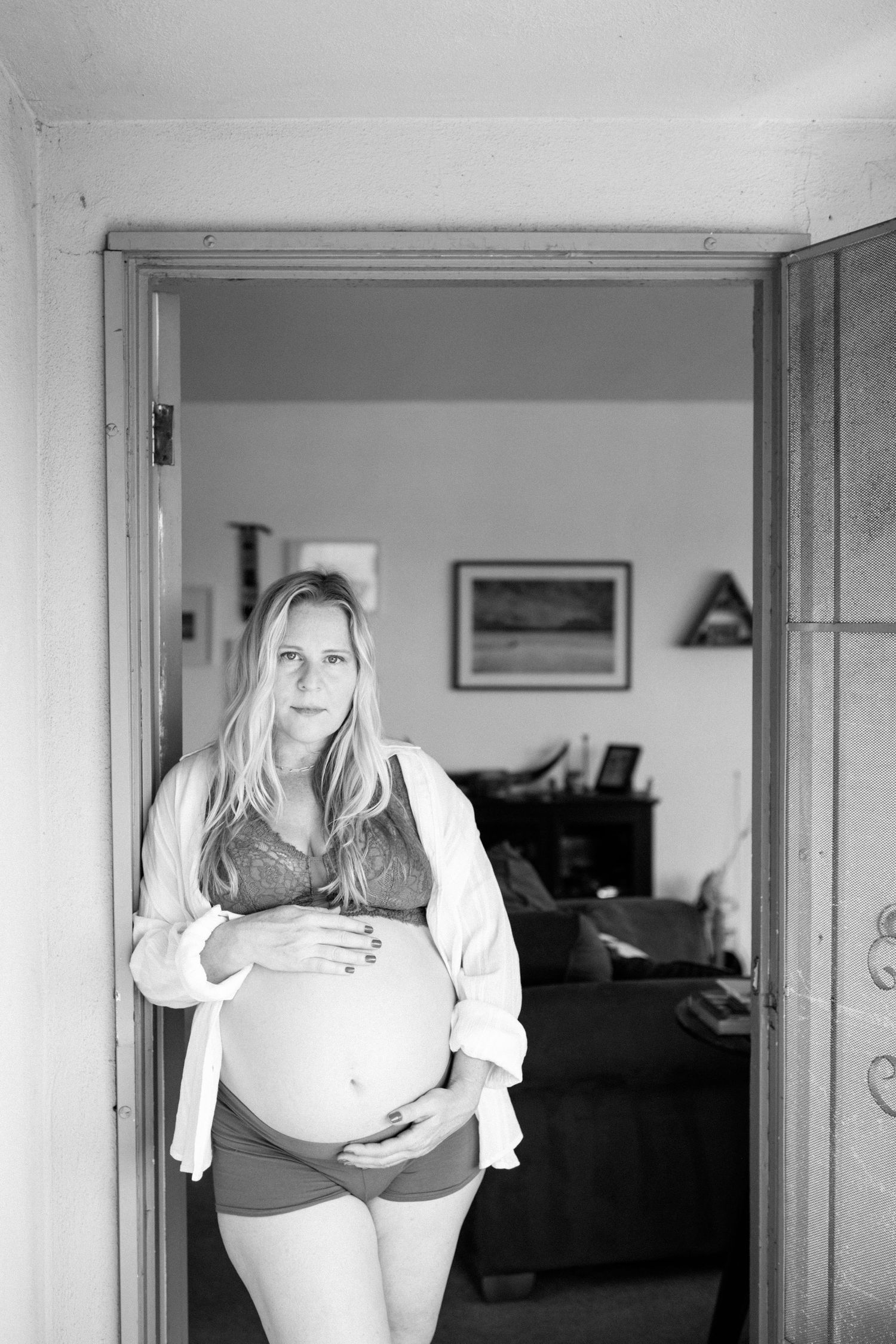 Blonde pregnant woman in open shirt and bra cradling bump in doorway, black and white.