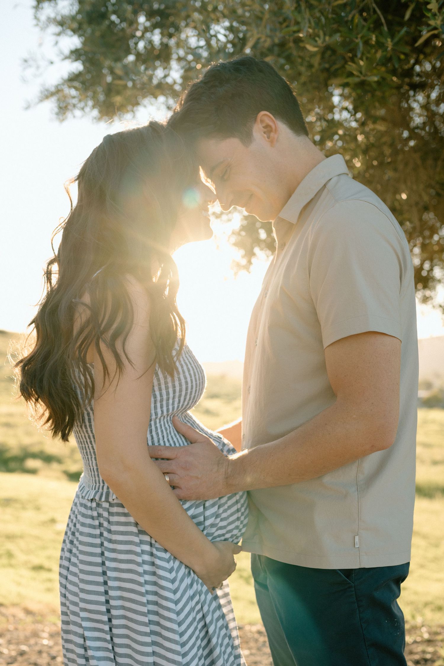 Couple stands forehead-to-forehead in sunlit park, man gently holds pregnant womans belly.
