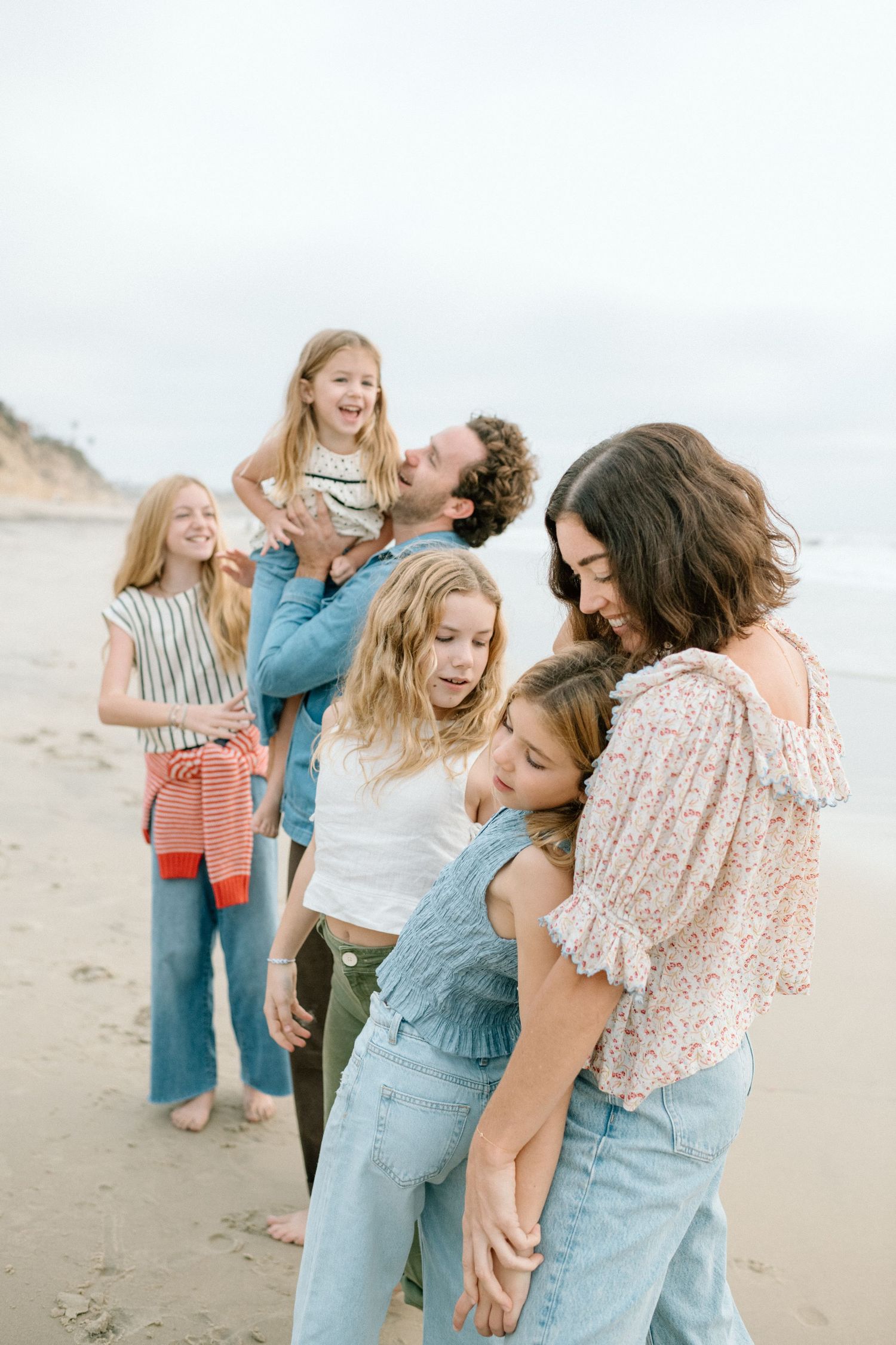 A happy family of six laughing and embracing on a sandy beach, with ocean cliffs visible in the background.