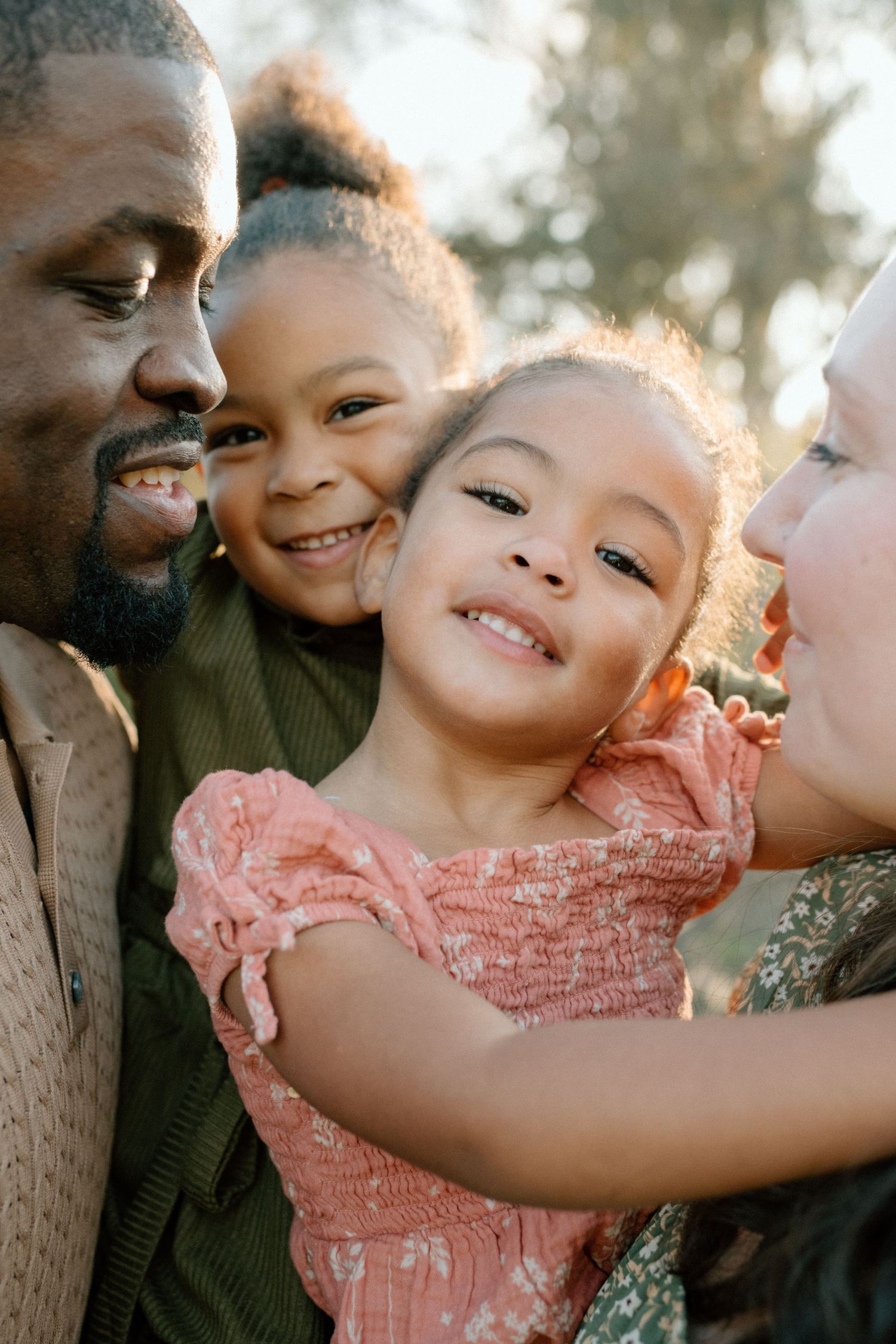 Joyful multiracial family with two young girls smiling closely together outdoors in soft natural light.