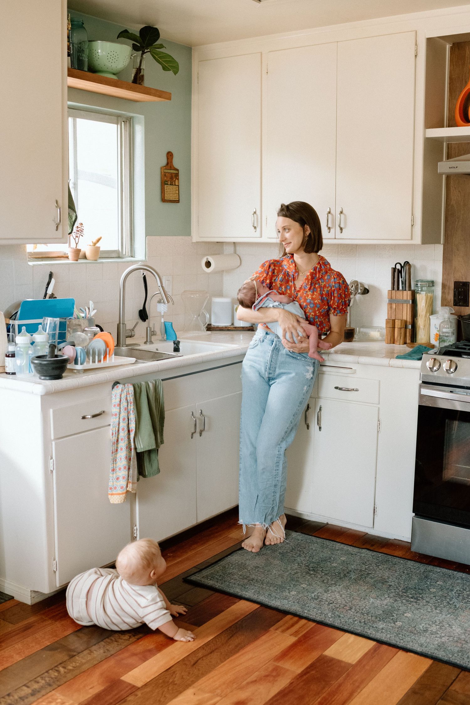 Mother holding newborn in white kitchen while baby crawls on hardwood floor nearby.