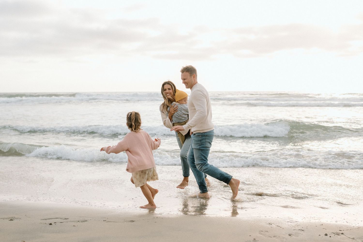 Family walking barefoot on a sandy beach near ocean waves, parents with two young children enjoying a seaside outing.
