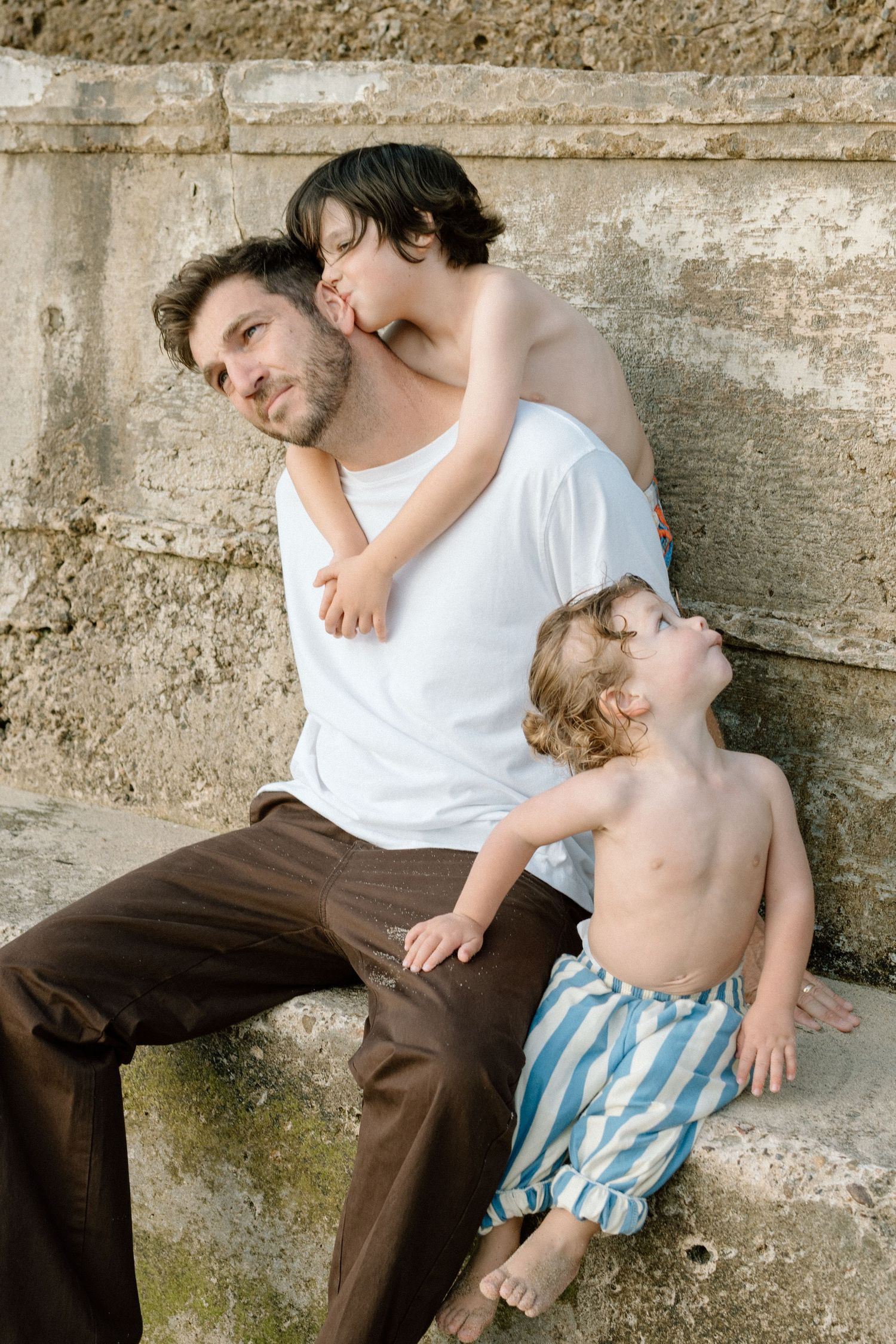 Father in white shirt sits against stone wall with two young children embracing him outdoors.