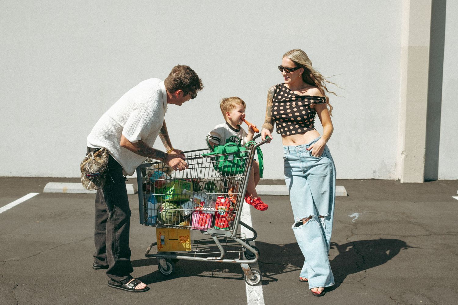 Family with toddler in shopping cart in parking lot, parents smiling in casual trendy outfits on sunny day.