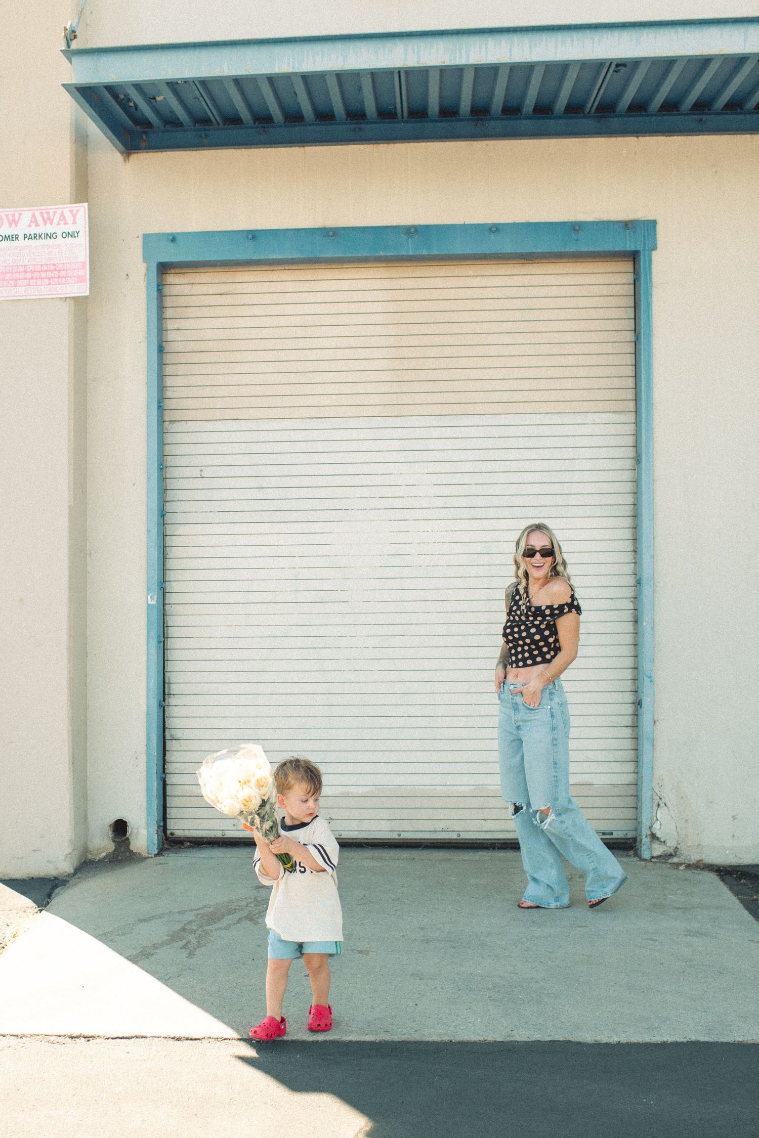 A woman in jeans and polka dot top stands near a garage door as a toddler holds white flowers outside a building.