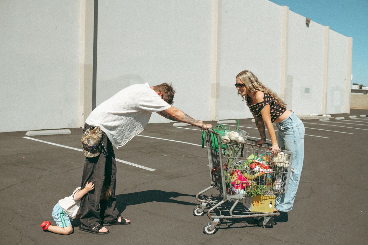 Two people with a shopping cart full of flowers in a parking lot, while a child plays near their feet.