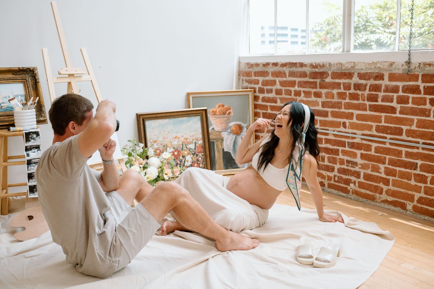 Sitting man takes picture of pregnant wife during maternity photoshoot in a San Diego studio.