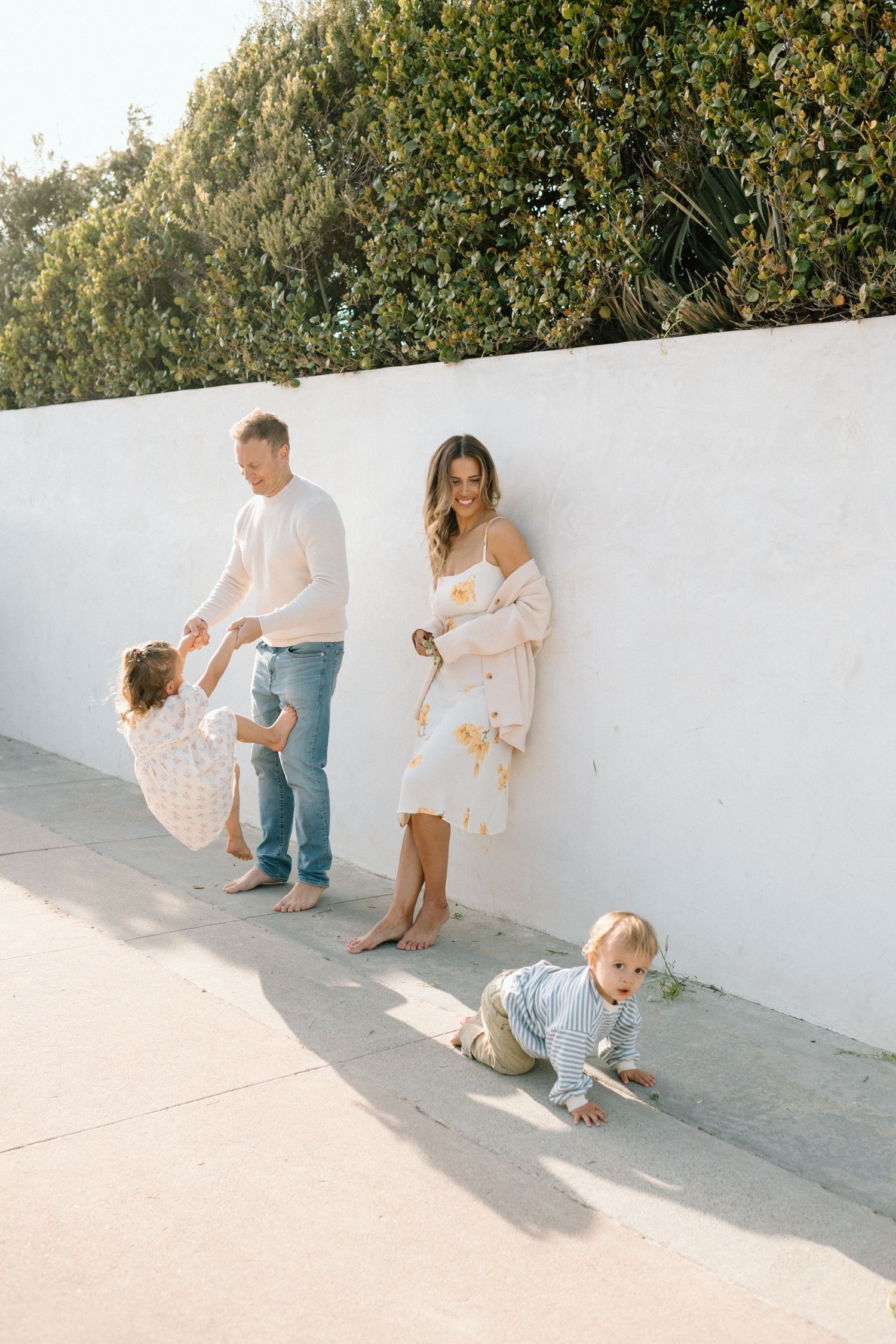 A barefoot family poses against a white wall outdoors, with dad twirling a toddler and a baby crawling nearby.