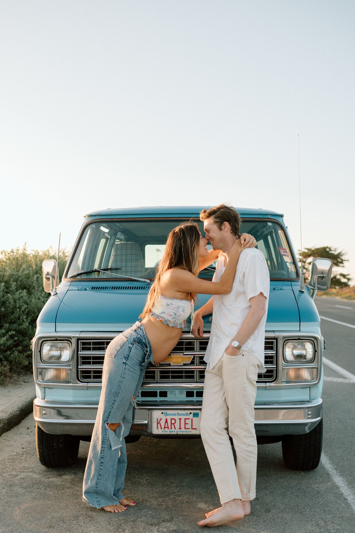 Couple sharing a romantic moment leaning against a classic blue van at golden hour.