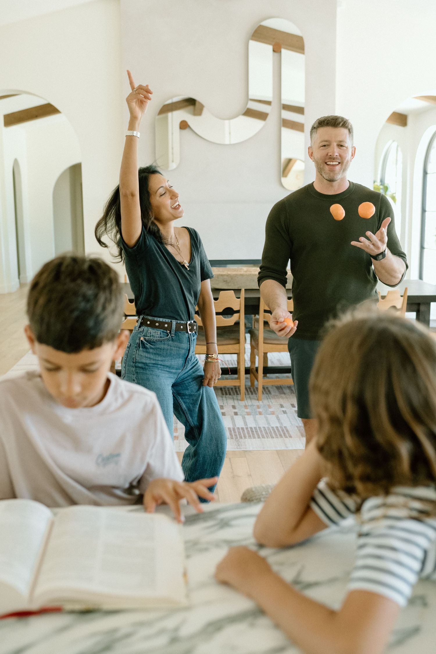 Parents dancing and juggling oranges while children read books at marble kitchen counter.