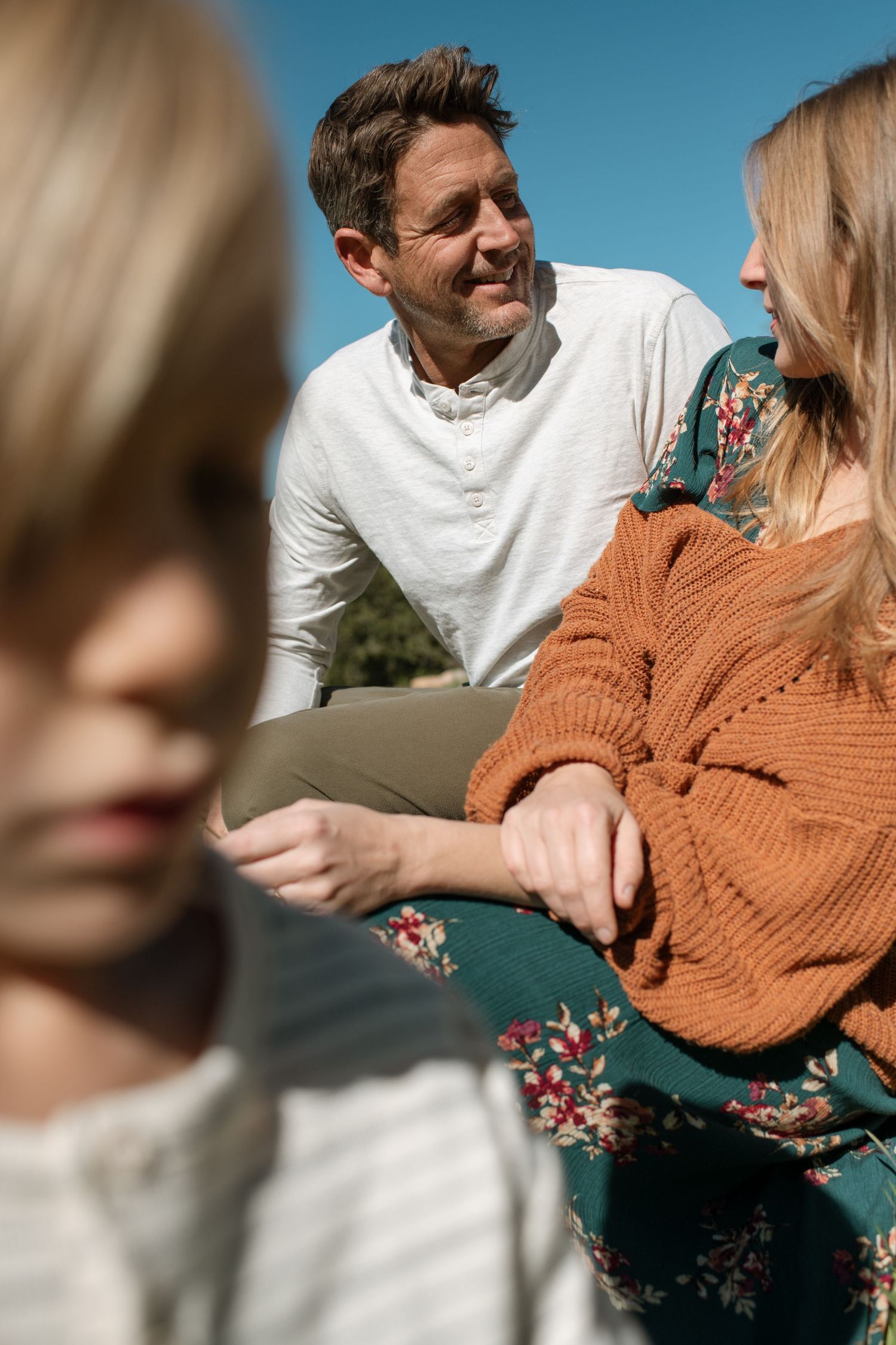 A smiling couple shares a tender moment outdoors while a blonde child stands in the foreground.