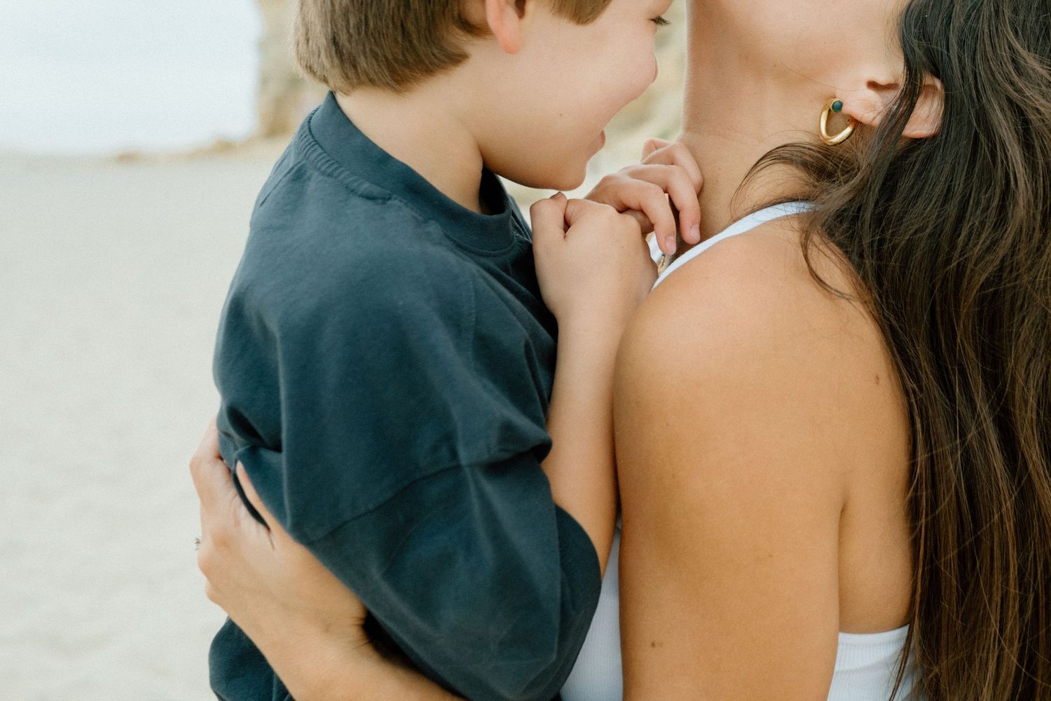 A young boy rests in his mothers arms on a sandy beach in a close tender moment.