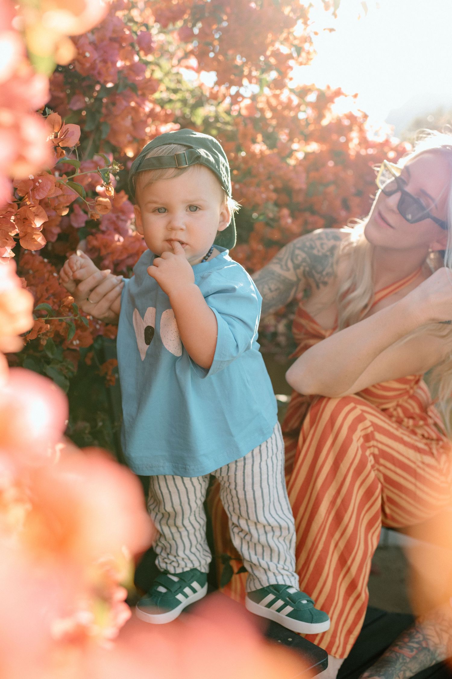 Toddler in blue tee and striped pants stands by pink bougainvillea flowers with mom nearby.