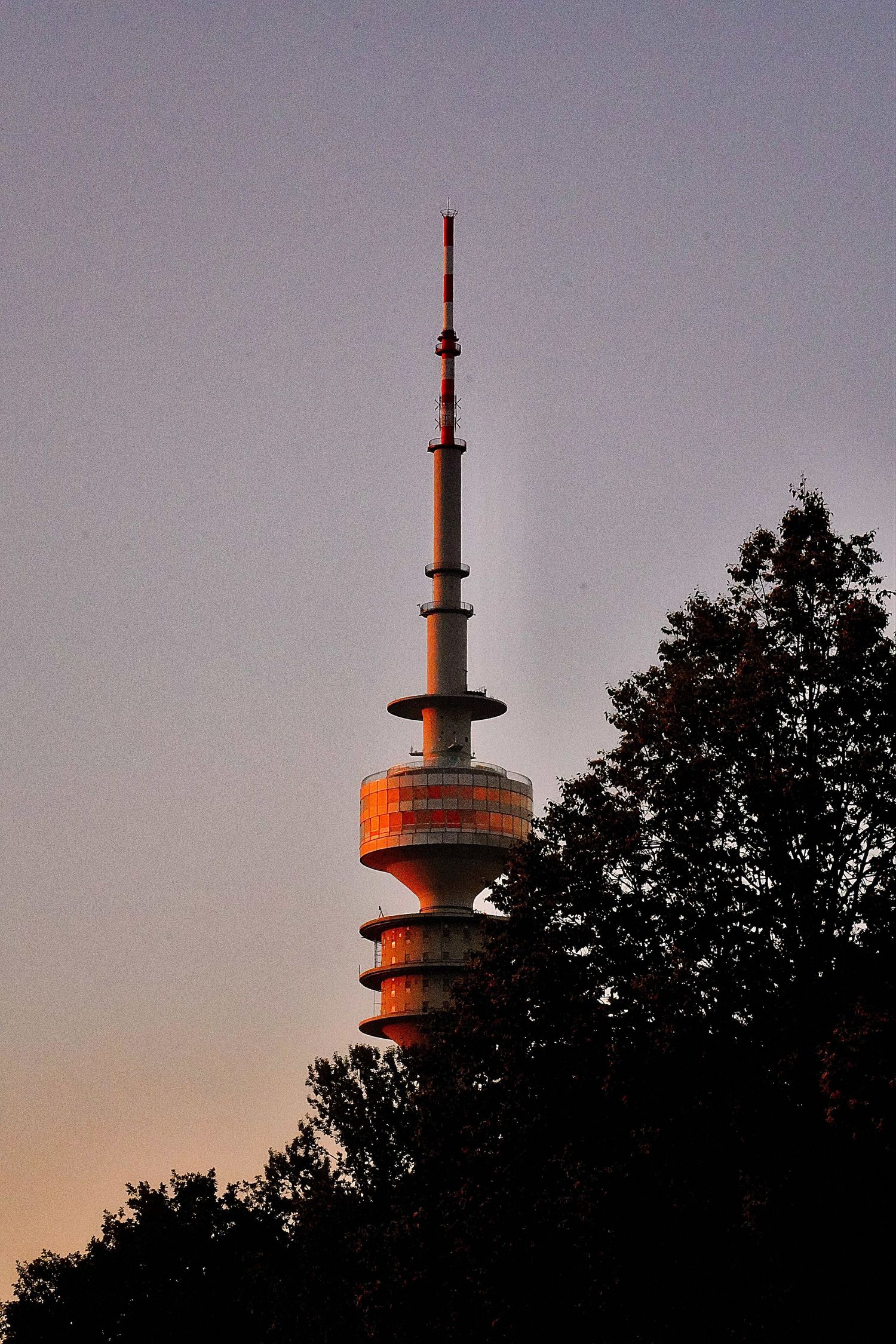 Olympic Tower in Munich bathed in warm sunset light, surrounded by dark tree silhouettes.