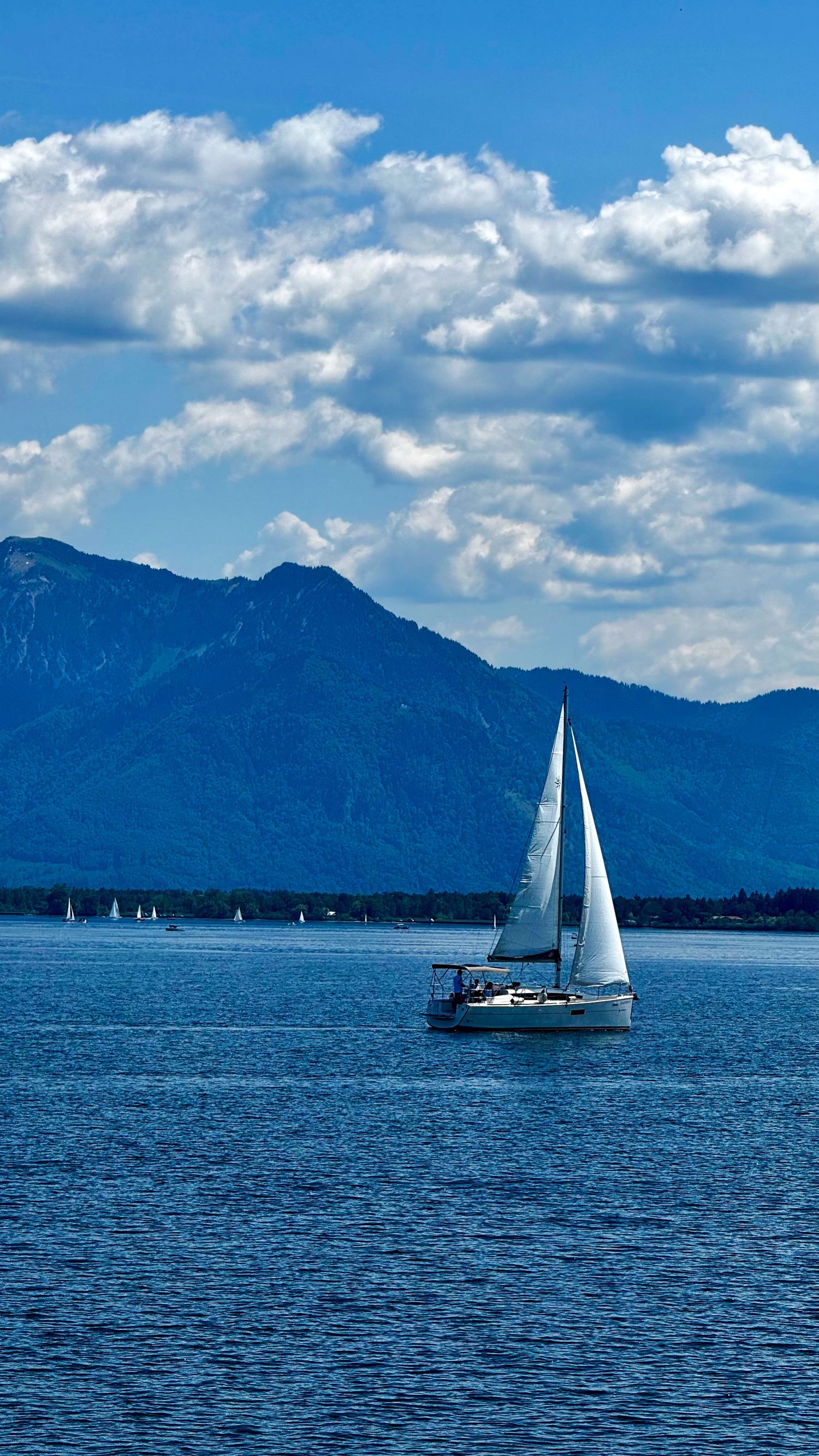 White sailboat gliding on blue lake with mountain backdrop and dramatic cloudy sky.