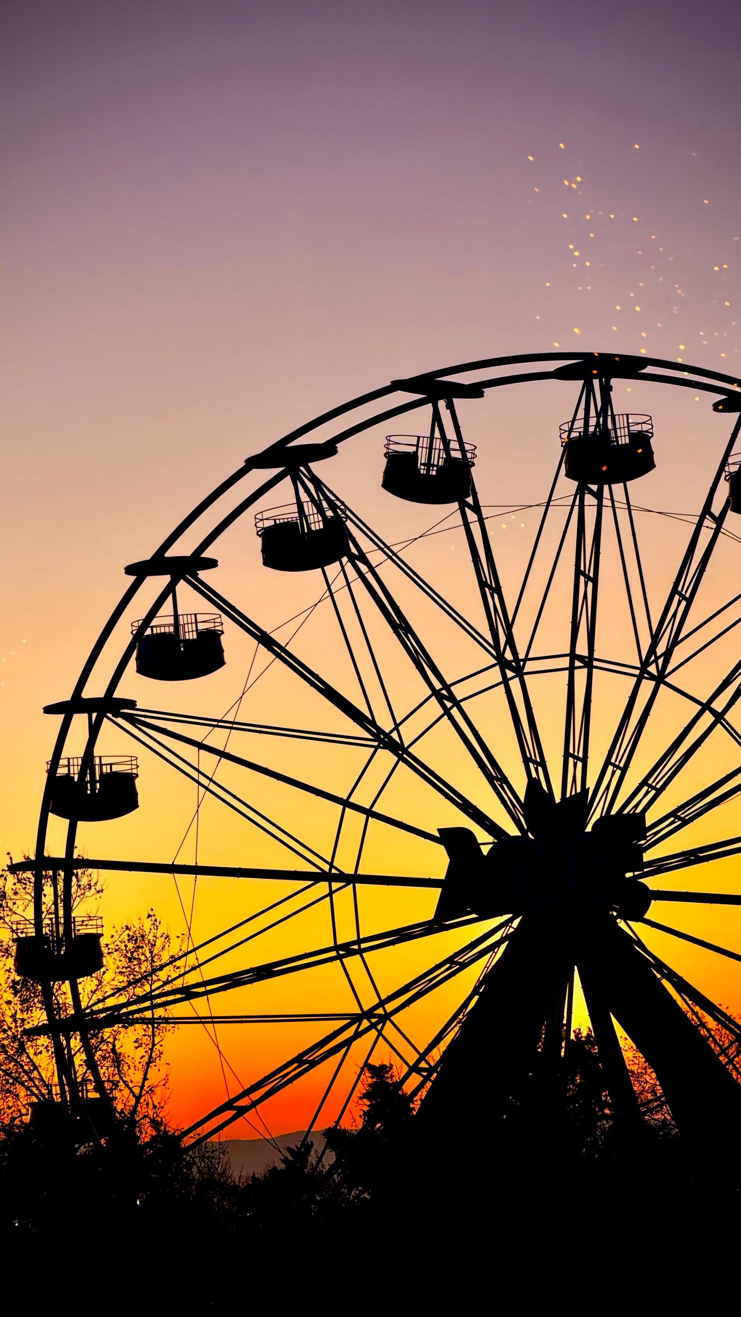 Silhouette of a ferris wheel against a vibrant orange and purple sunset sky with sparkling lights.