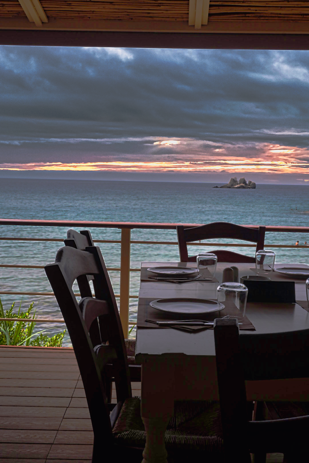 Oceanfront restaurant terrace with set dining table overlooking dramatic sunset over sea.