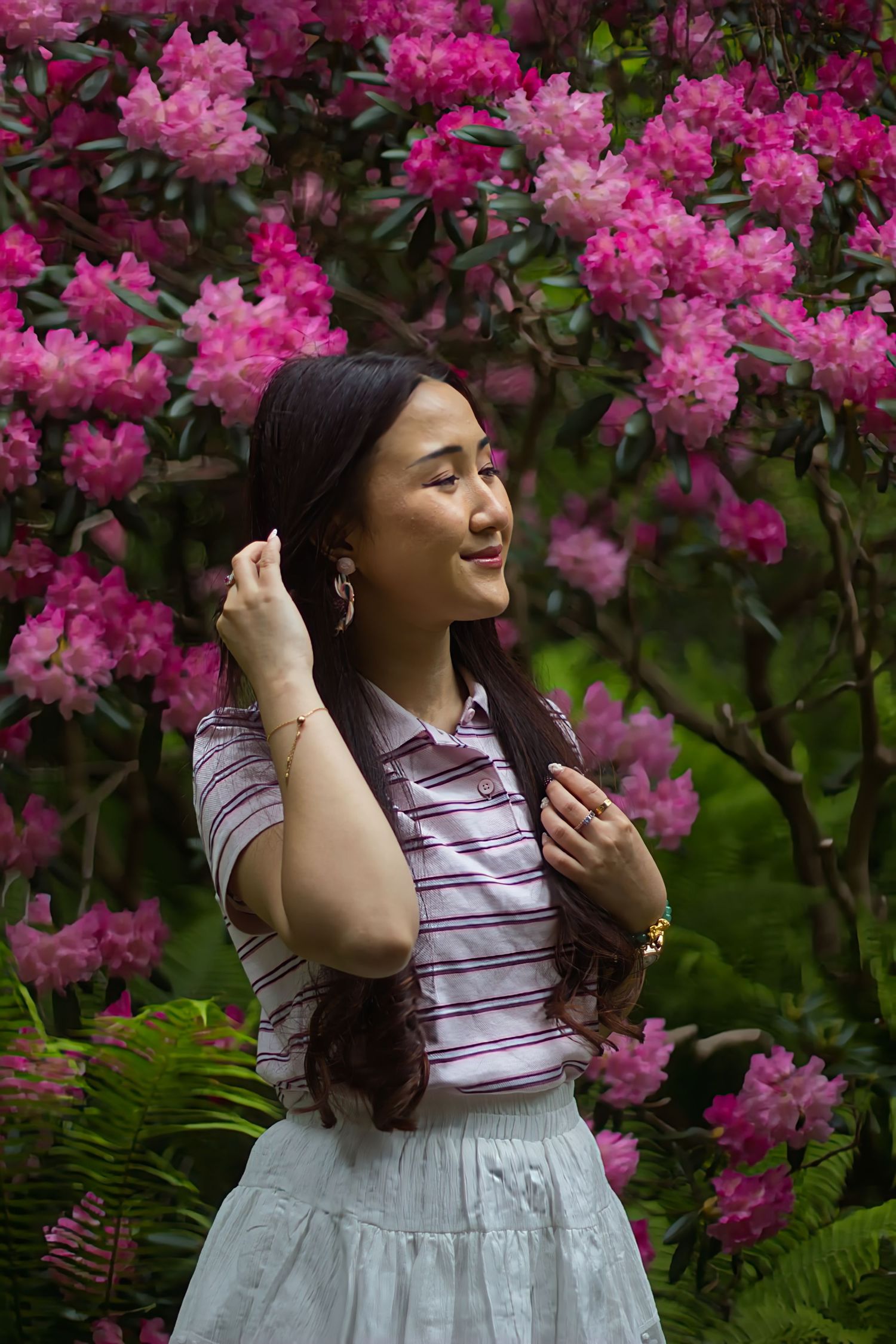 Smiling Asian woman in striped shirt posing before vibrant pink rhododendron blooms.